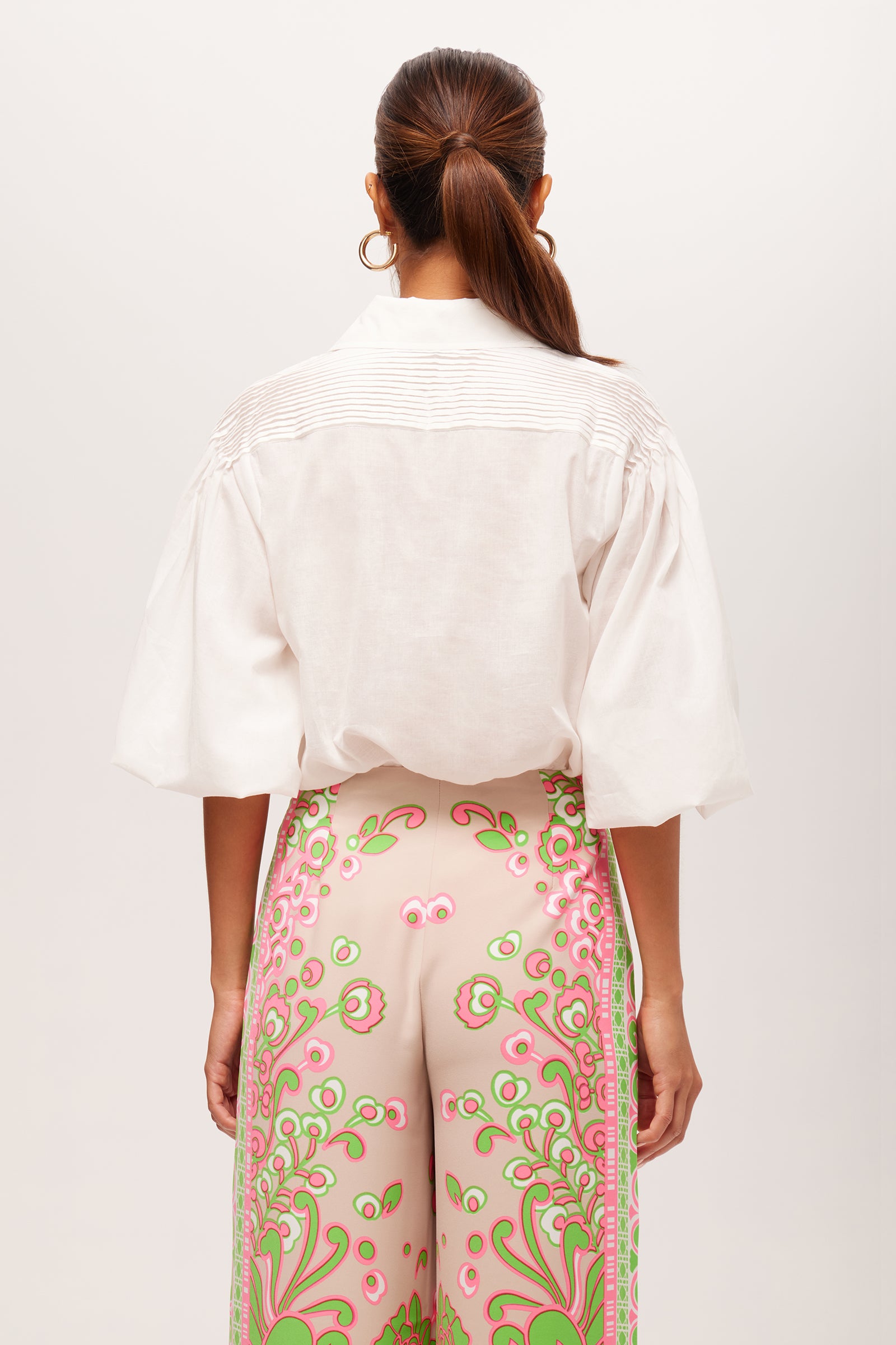 A woman with long brown hair in a ponytail is shown from the back, wearing the Court Short-Sleeve Button-Up Top in white and high-waisted beige pants with a bold pink and green floral pattern.