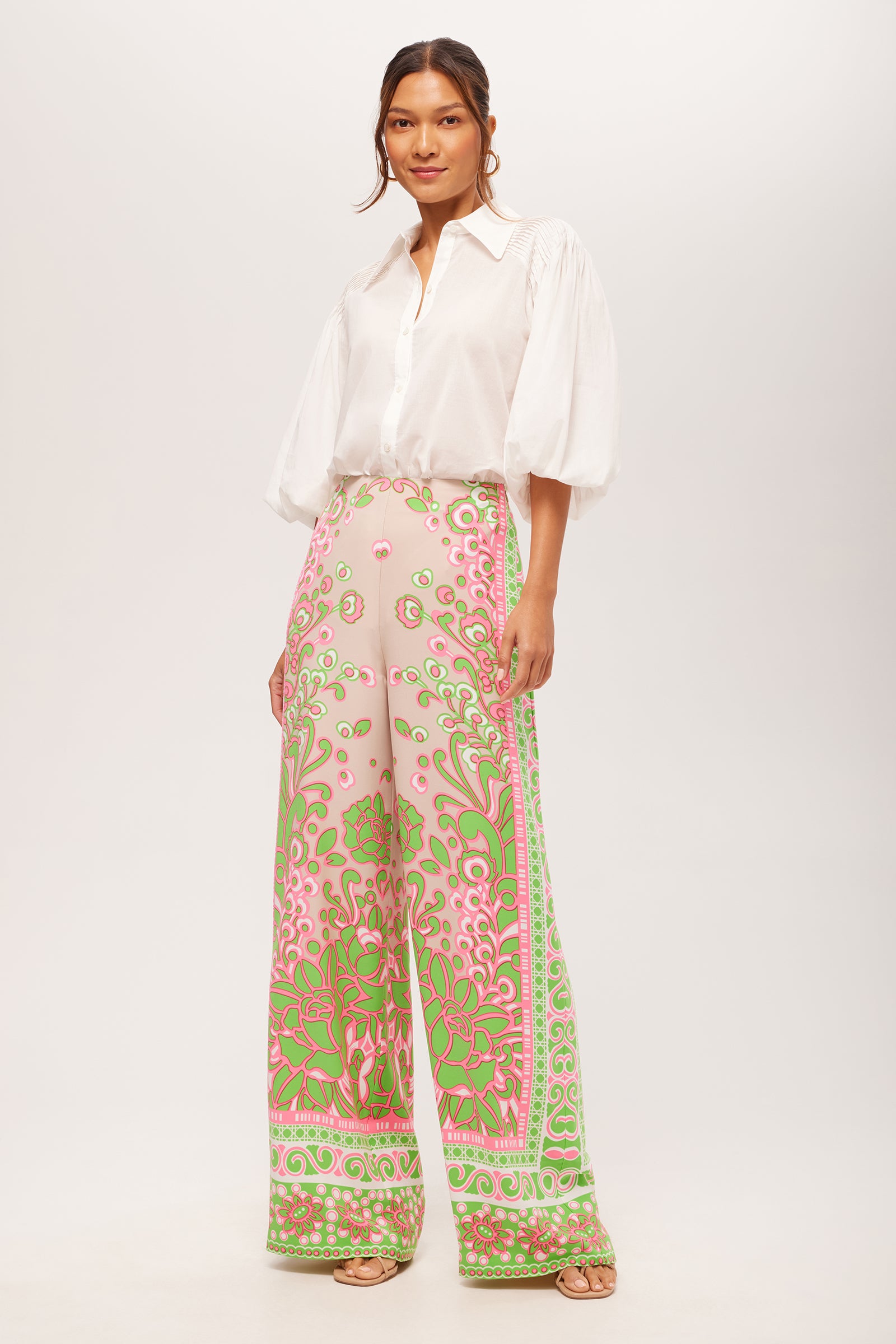 A woman with long hair tied back wears the Court Short-Sleeve Button-Up Top and high-waisted, wide-leg pants with a pink, green, and gold ornate floral pattern. She stands against a plain background and pairs the look with sandals.