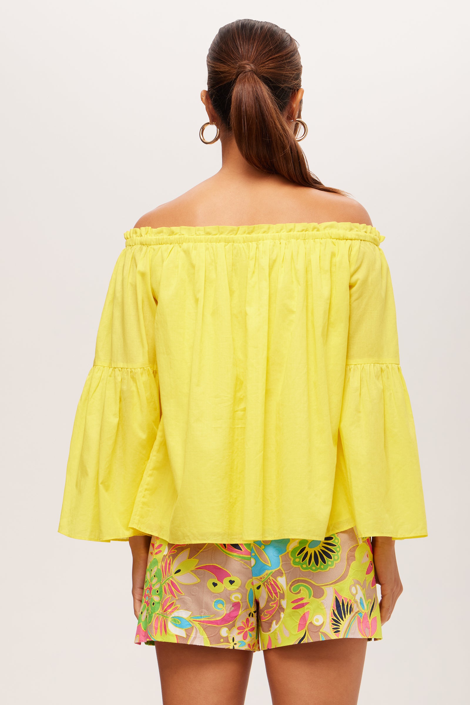 A woman faces away, wearing the Perry Off-Shoulder Long-Sleeve Top in bright yellow with bell sleeves and colorful floral shorts. Her hair is in a low ponytail, she wears gold hoop earrings, and the background is plain and light.