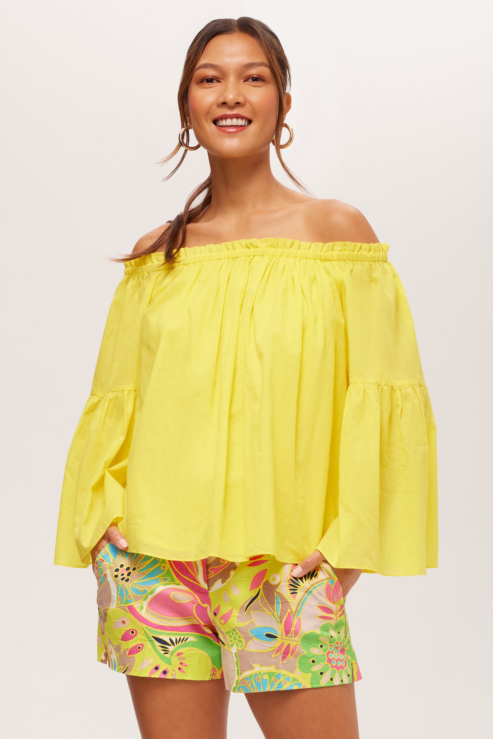 A smiling woman stands with hands in her shorts’ pockets, wearing the Perry Off-Shoulder Long-Sleeve Top in Desert Sun and colorful floral shorts. Her hair is in a loose ponytail, complemented by hoop earrings.