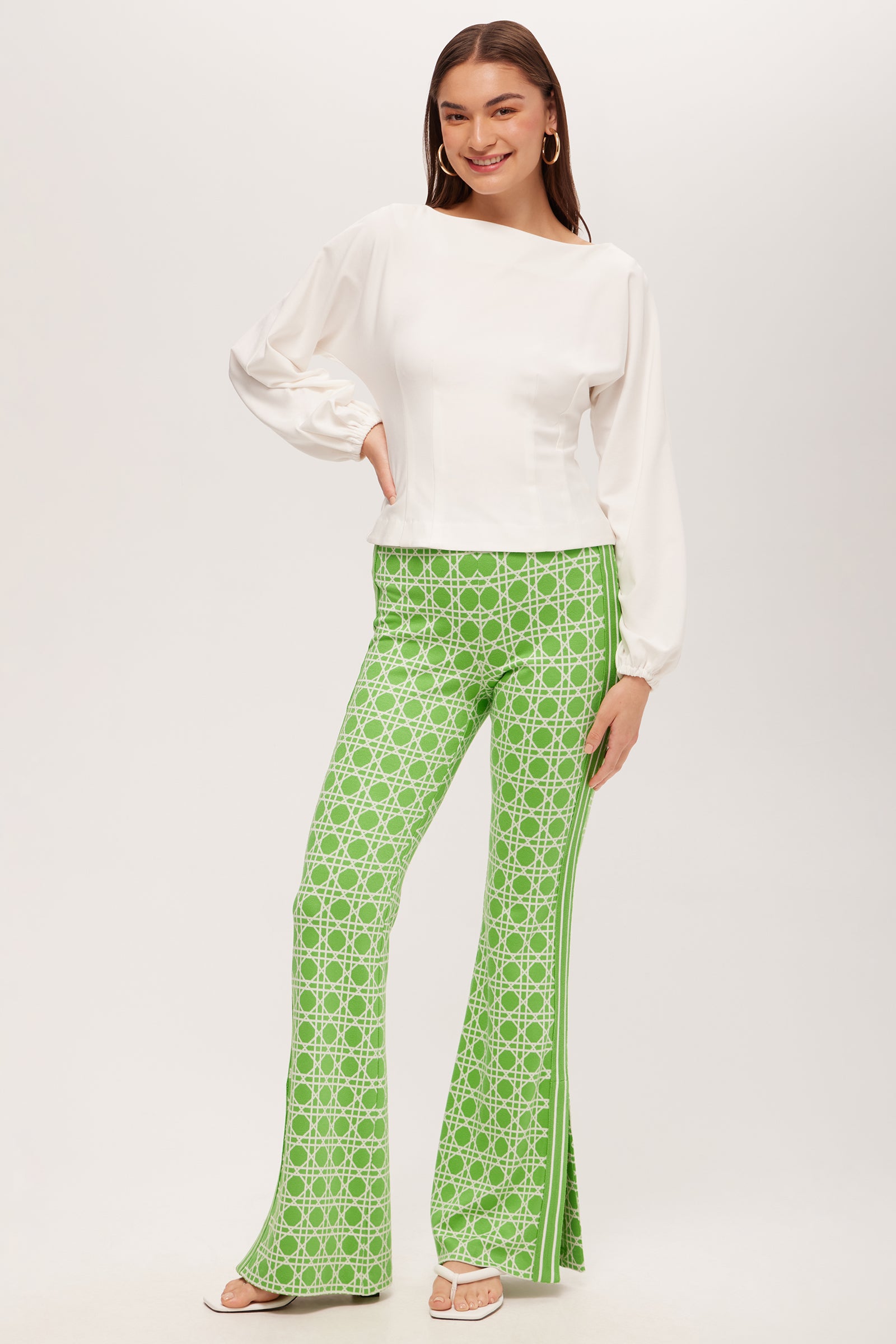 A woman with long brown hair smiles, wearing the Prime Long-Sleeve Boatneck Top - Cozy Ponte in white and high-waisted green patterned flared pants with side slits, paired with sandals, against a plain white background.