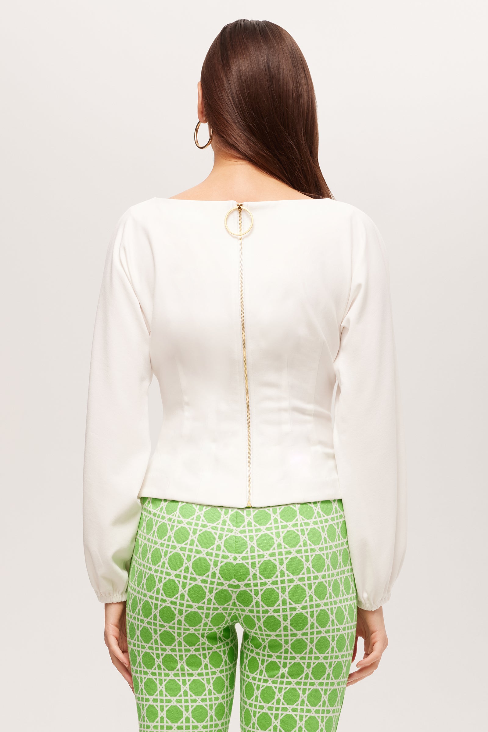 A woman faces away from the camera, wearing the Prime Long-Sleeve Boatneck Top - Cozy Ponte in white with a gold zipper and ring pull, paired with green pants featuring a white geometric pattern.
