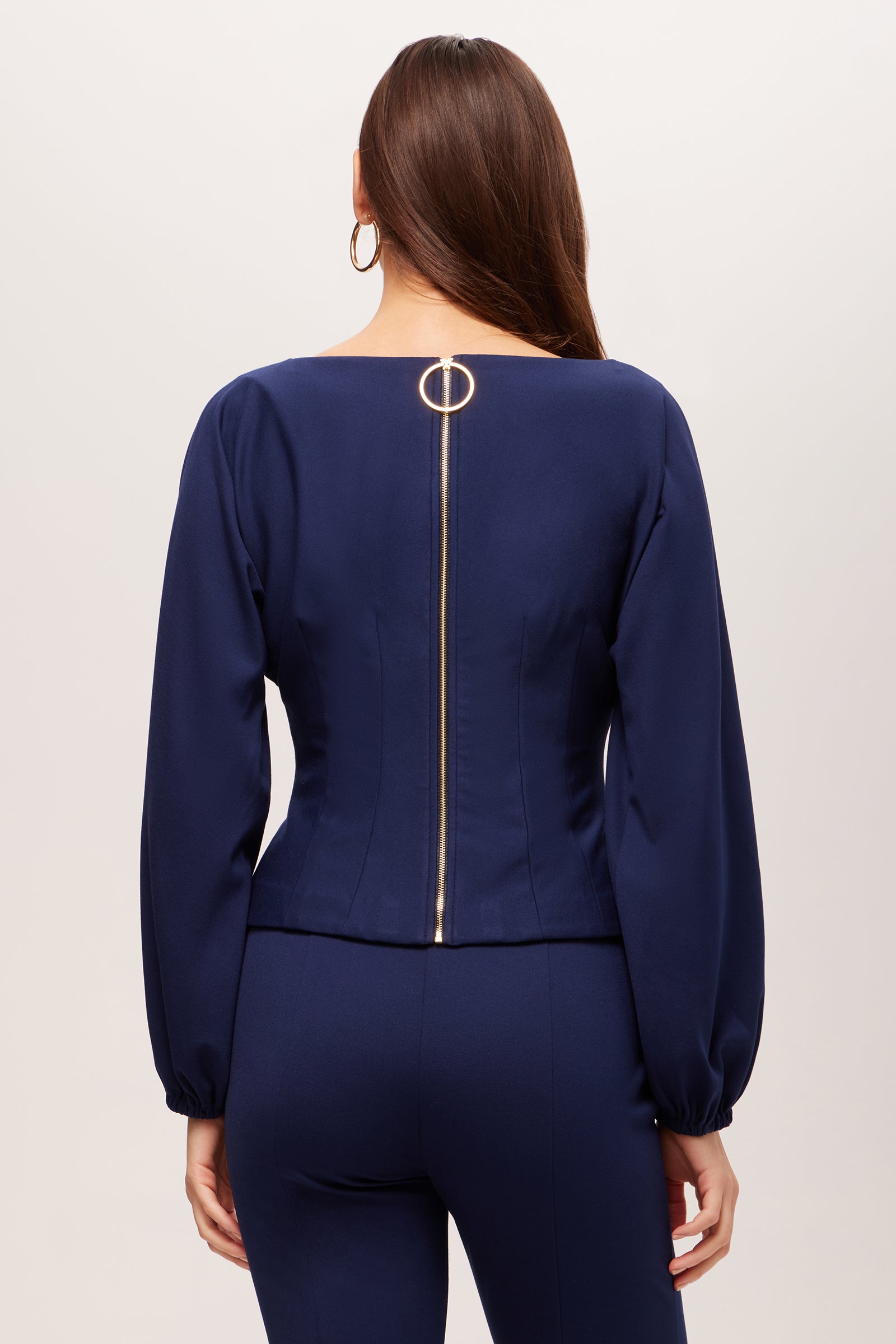 A woman with long brown hair is seen from the back wearing the Prime Long-Sleeve Boatneck Top - Cozy Ponte in navy blue, featuring a gold center zipper with a circular pull, paired with matching navy blue pants.