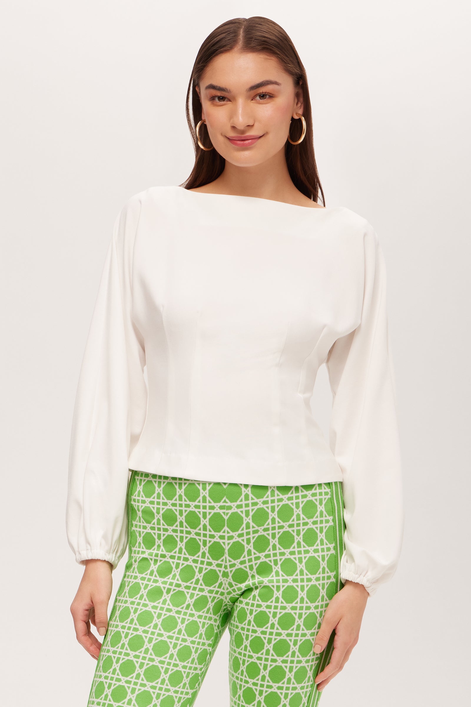 A woman with straight brown hair and a slight smile stands against a plain background, wearing the Prime Long-Sleeve Boatneck Top in Whitewash and green pants with a white geometric pattern.