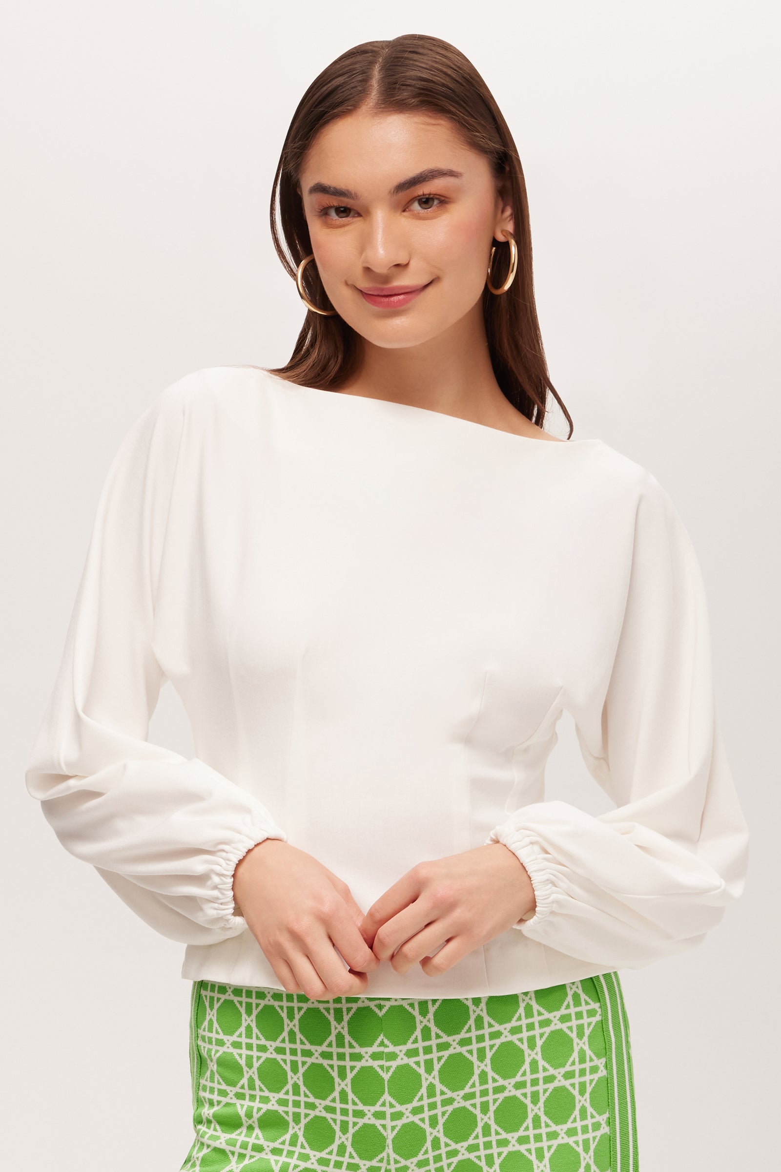 A woman with long brown hair wears the Prime Long-Sleeve Boatneck Top - Cozy Ponte in white, paired with a green skirt featuring a white geometric pattern. She stands against a light background, smiling softly.