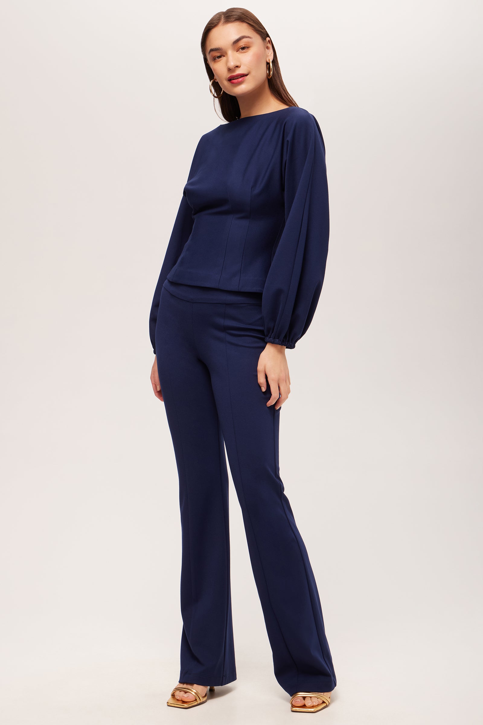A woman poses against a plain background wearing the Becker High-Waist Flared Pant - Cozy Ponte in navy blue, paired with a matching long-sleeve top, gold sandals, hoop earrings, and styled with long, straight hair.