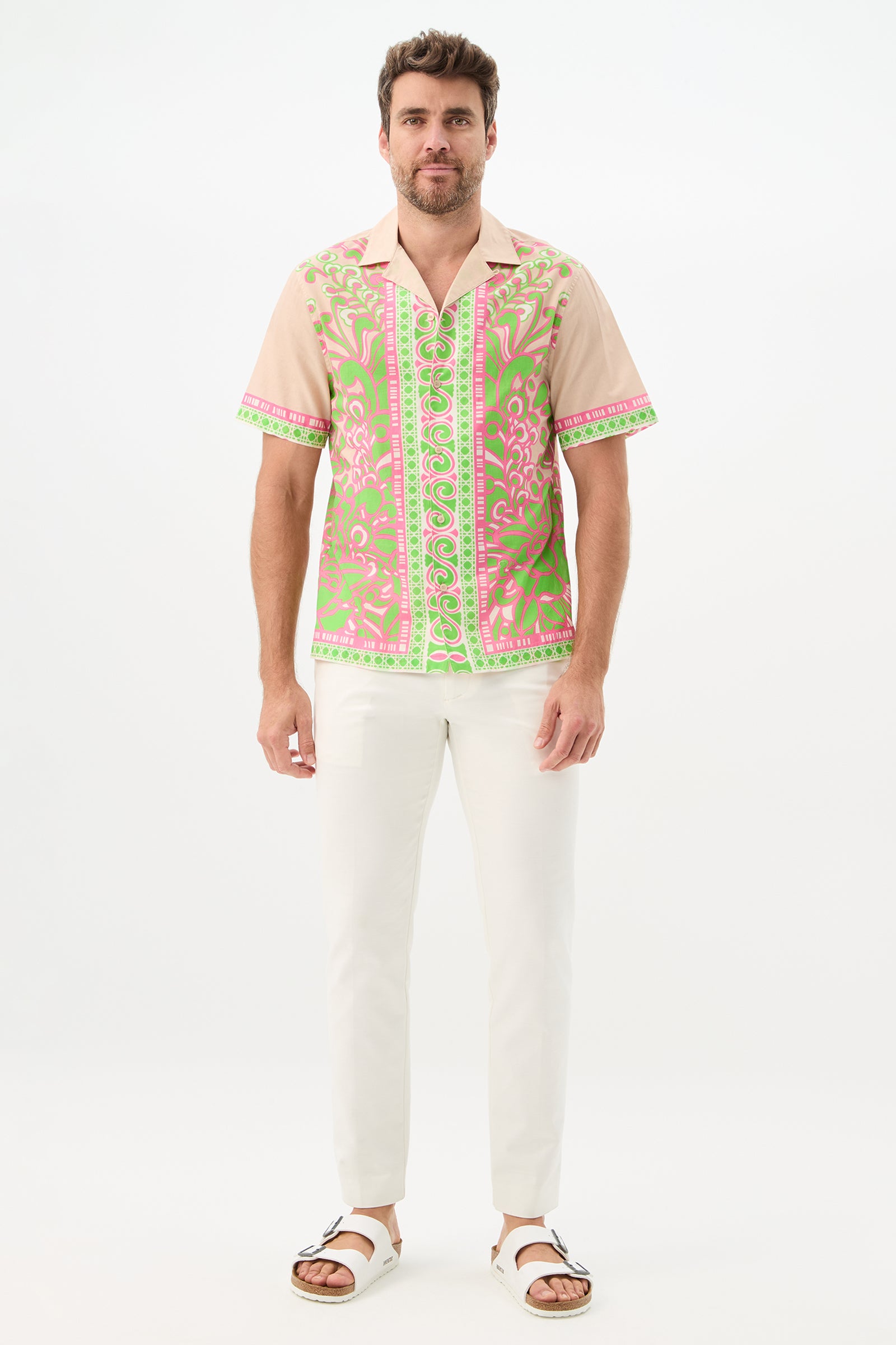 A man with short brown hair and a beard stands against a white background, smiling slightly in the Josue Short-Sleeve Shirt - Petra Floral, featuring a pink and green ornate pattern, paired with white pants and sandals.