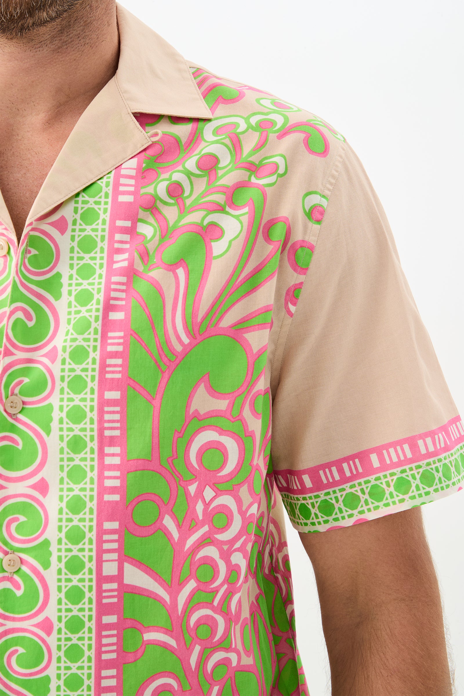 A man wearing the Josue Short-Sleeve Shirt - Petra Floral, featuring vibrant green and pink swirling patterns and geometric designs on beige. Only his upper torso and part of his face are visible against a plain white background.