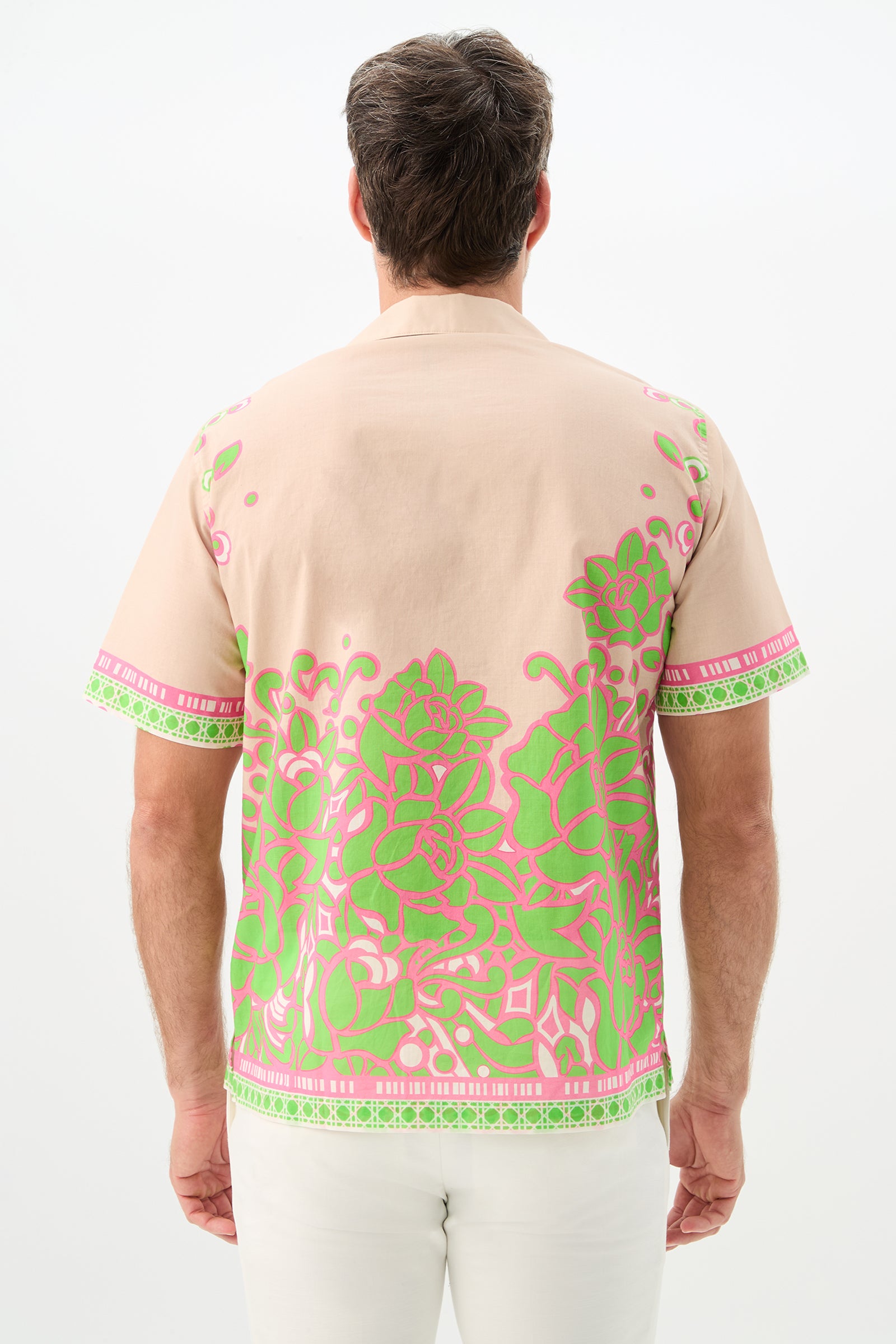 A man is seen from behind wearing the Josue Short-Sleeve Shirt - Petra Floral, featuring bright green and pink floral patterns on beige, and white pants against a plain white background.