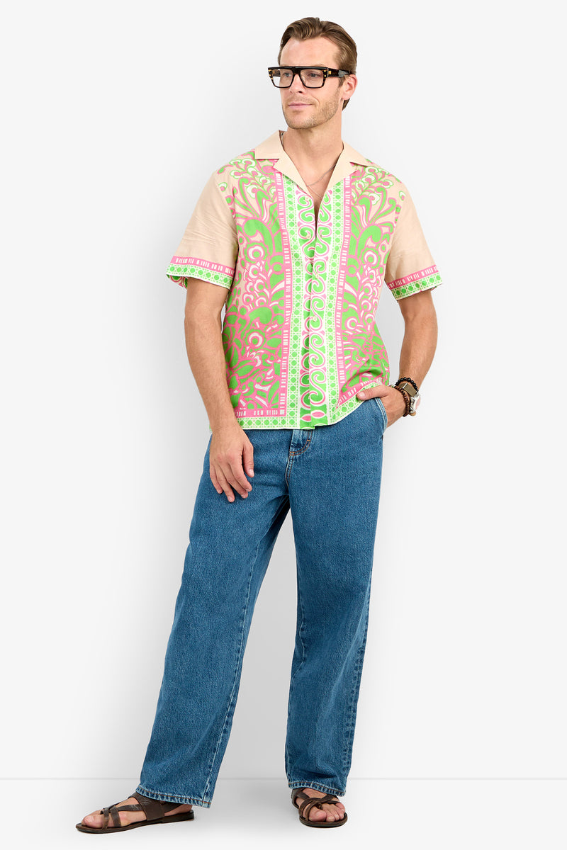 Man wearing glasses, the Josue Camp Shirt with pink and green patterns, blue jeans, sandals, and a watch stands with one hand in his pocket against a plain white background.