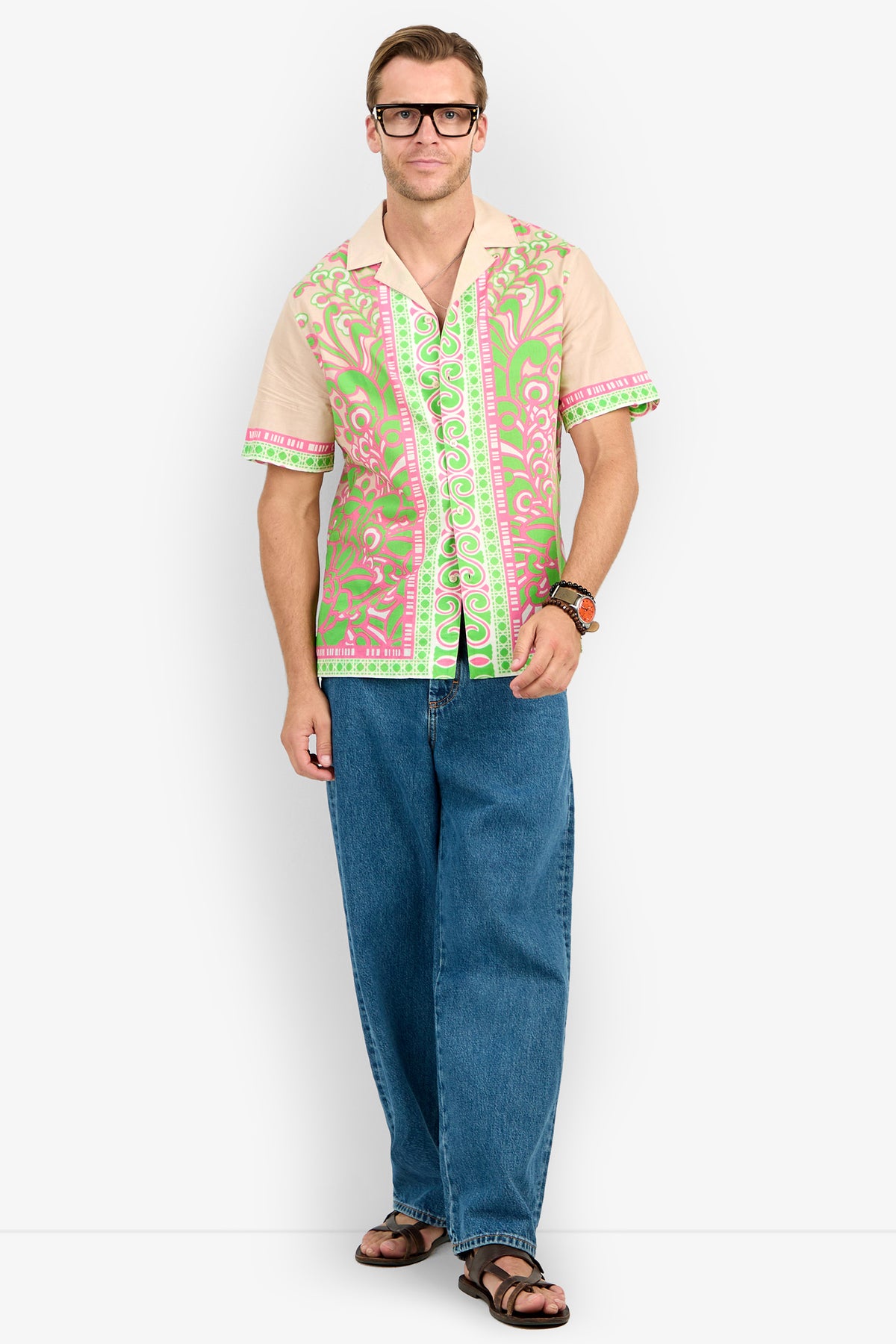 Man wearing black glasses, the Josue Camp Shirt with pink and green patterns, blue wide-leg jeans, and brown sandals stands against a plain white background.