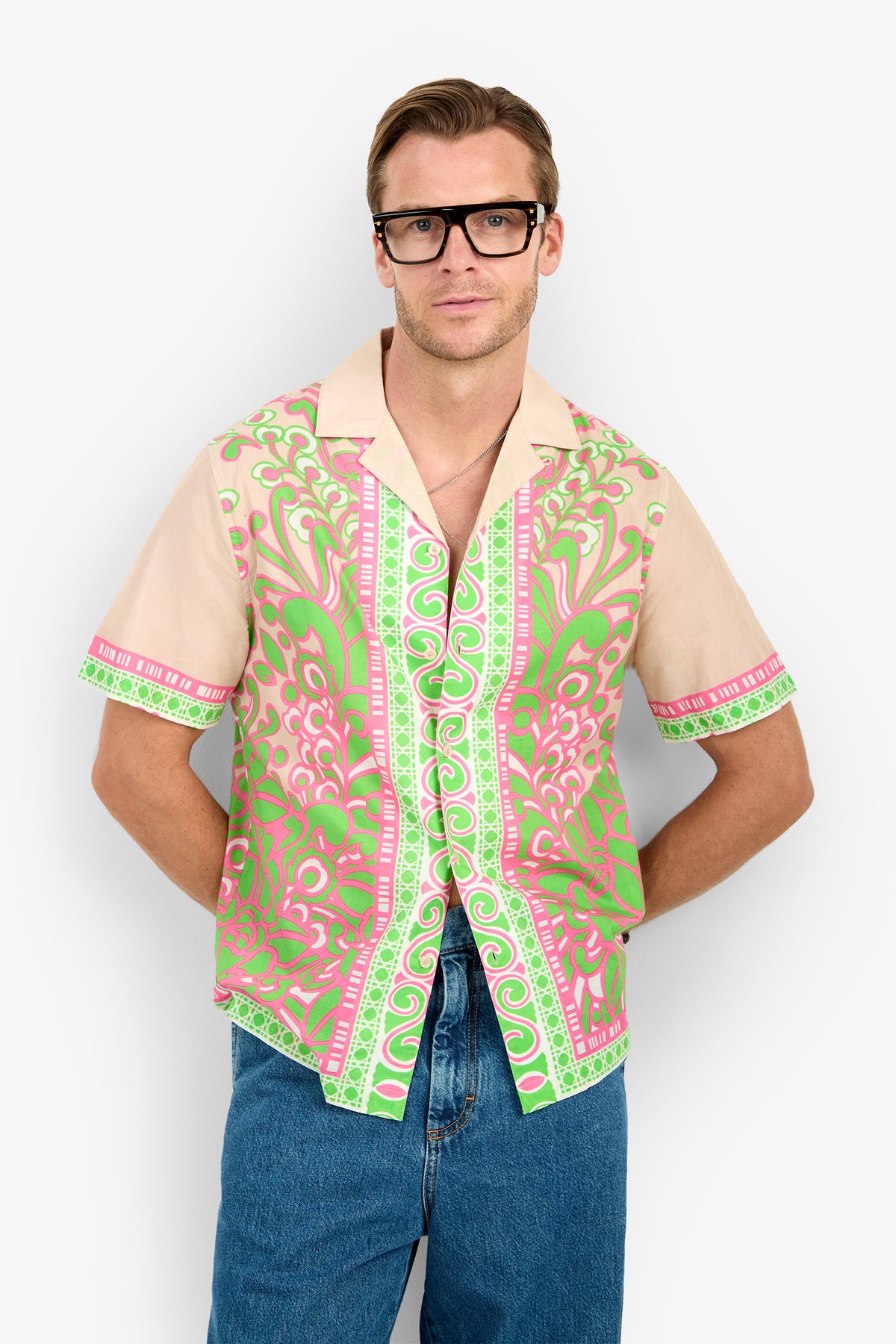 A man wearing glasses, blue jeans, and the Josue Camp Shirt with pink and green patterns stands against a plain white background with his hands in his pockets.