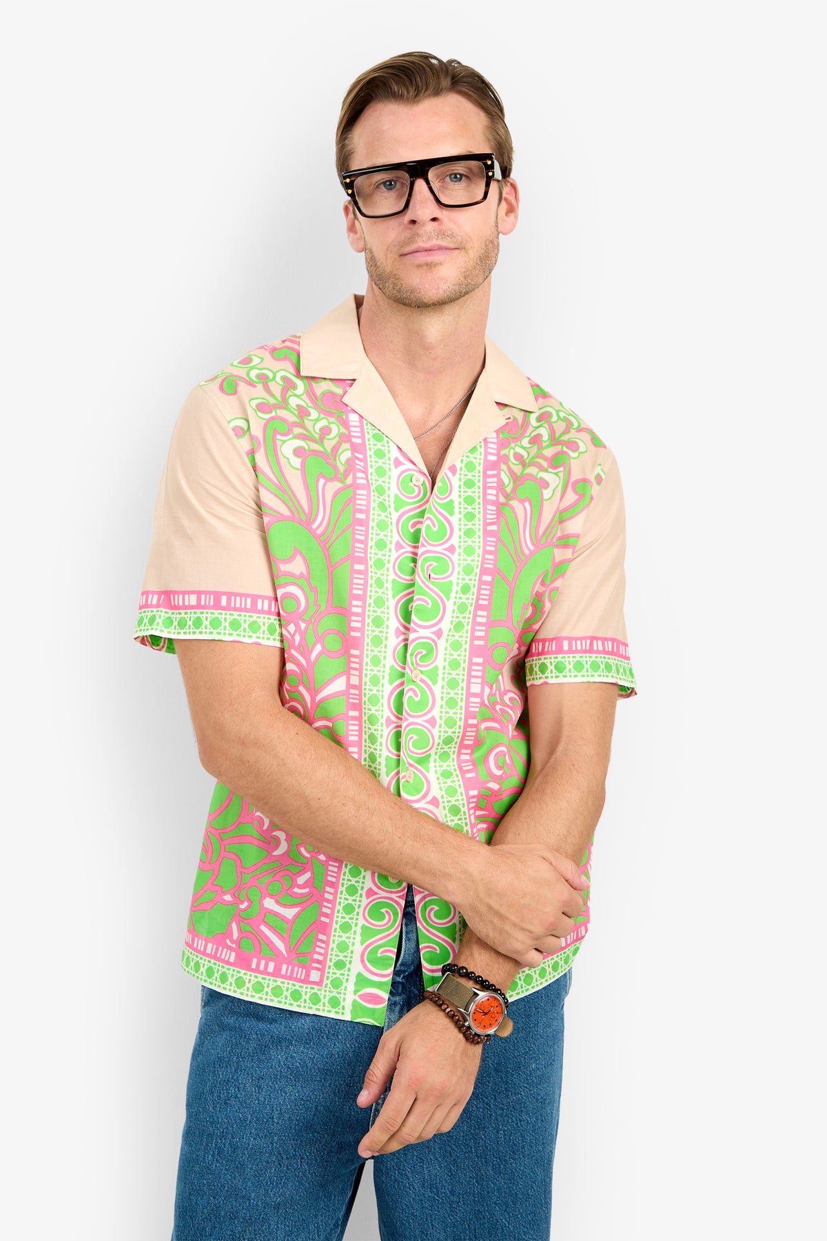 A man wearing black glasses, blue jeans, and the Josue Camp Shirt—a colorful short-sleeve shirt with pink and green patterns—stands with his arms crossed in front of a plain background.