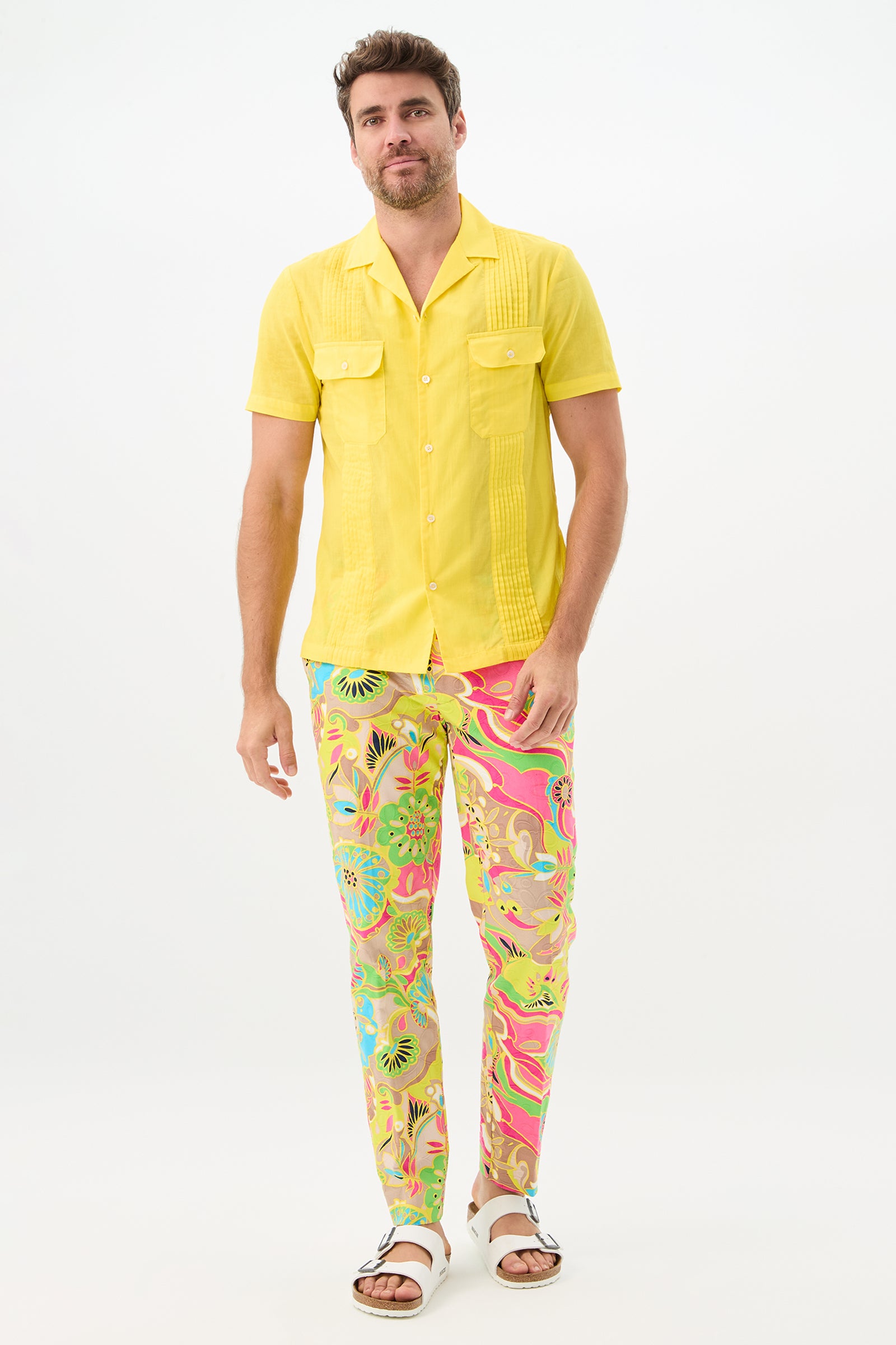 A man with short brown hair and a light beard stands against a white background, smiling slightly while wearing colorful Clyde Slim Trouser - Venus pants, a bright yellow short-sleeve shirt, and white sandals.