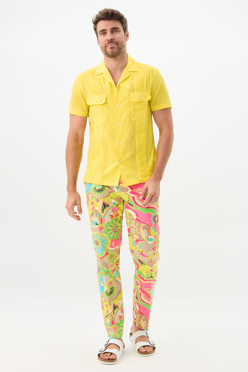 A man with short brown hair and a light beard stands against a white background, smiling slightly while wearing colorful Clyde Slim Trouser - Venus pants, a bright yellow short-sleeve shirt, and white sandals.
