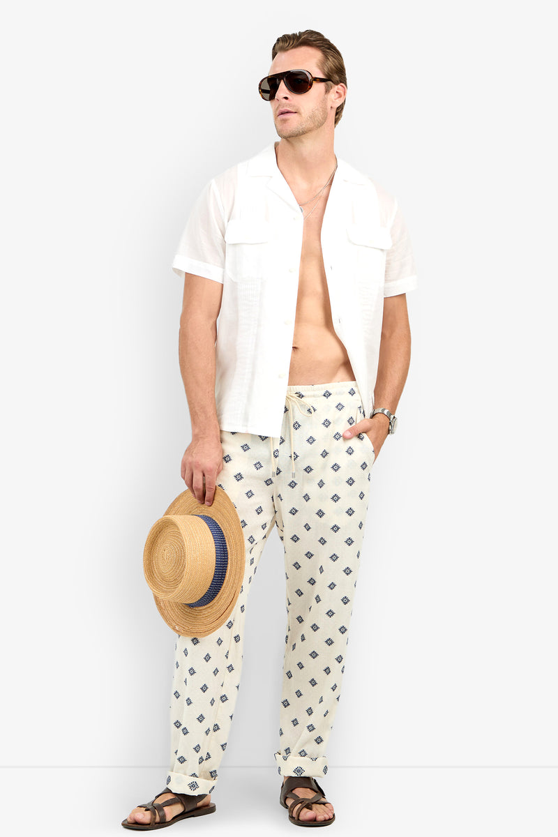 A man in the Allende Short-Sleeve Shirt, patterned white pants, sunglasses, and sandals stands holding a straw hat, looking to the side against a plain background.