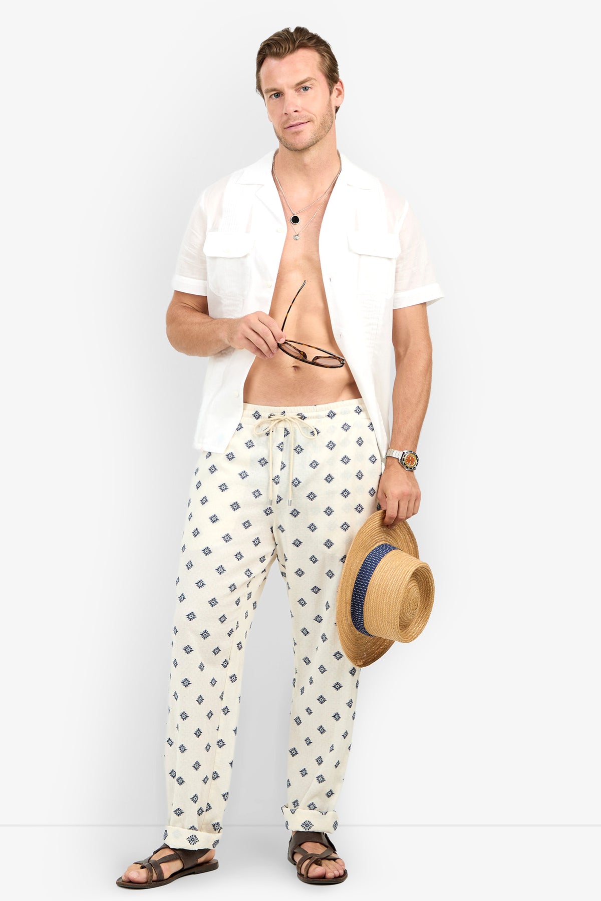 A man wears the Allende Short-Sleeve Shirt open over drawstring pants and sandals, holding sunglasses and a straw hat. He stands against a plain background, relaxed and stylish.
