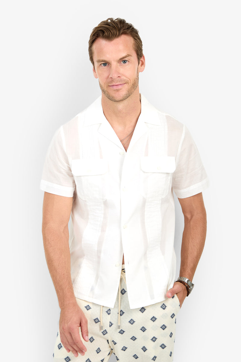 A man wearing the Allende Short-Sleeve Shirt in white with front pockets and patterned shorts stands against a plain light background, looking at the camera and smiling slightly.