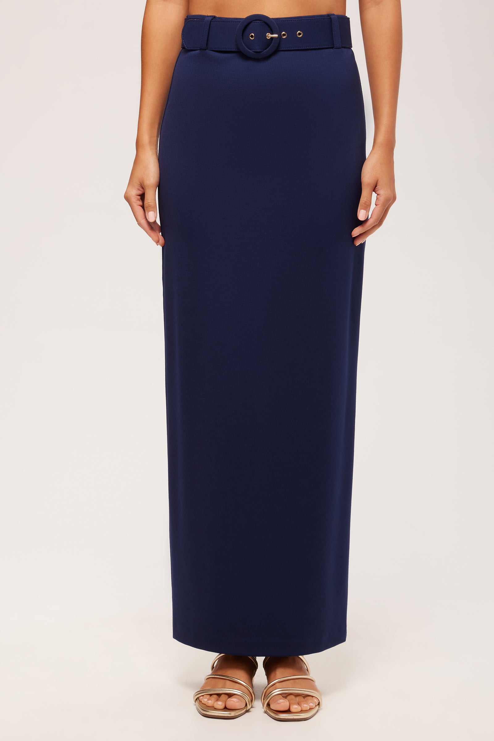 A person wears the Game Point Column Maxi Skirt - Cozy Ponte in Ink, featuring a high waist, wide belt with round buckle, paired with flat tan sandals, standing against a plain light background.