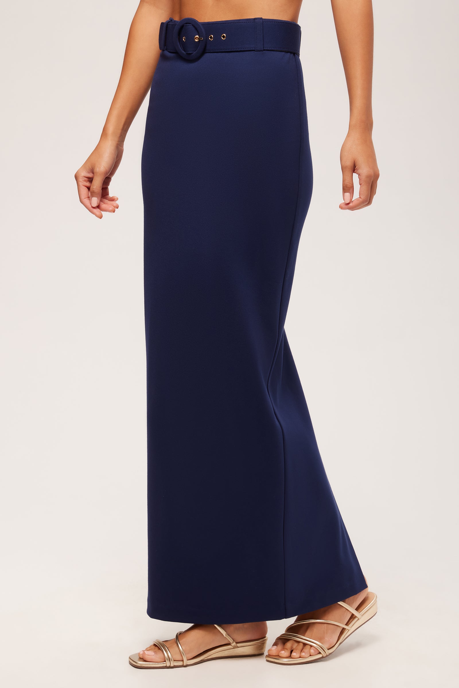 A person wearing the Game Point Column Maxi Skirt - Cozy Ponte in navy blue with a matching belt and gold sandals, standing against a plain light background.