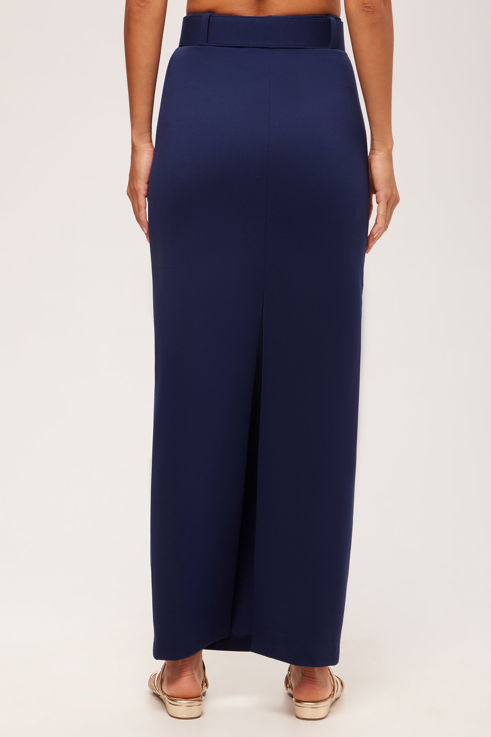 A person wearing the Game Point Column Maxi Skirt - Cozy Ponte, a high-waisted, floor-length navy blue skirt with a back slit, paired with beige open-toe sandals, shown from the back against a plain light background.