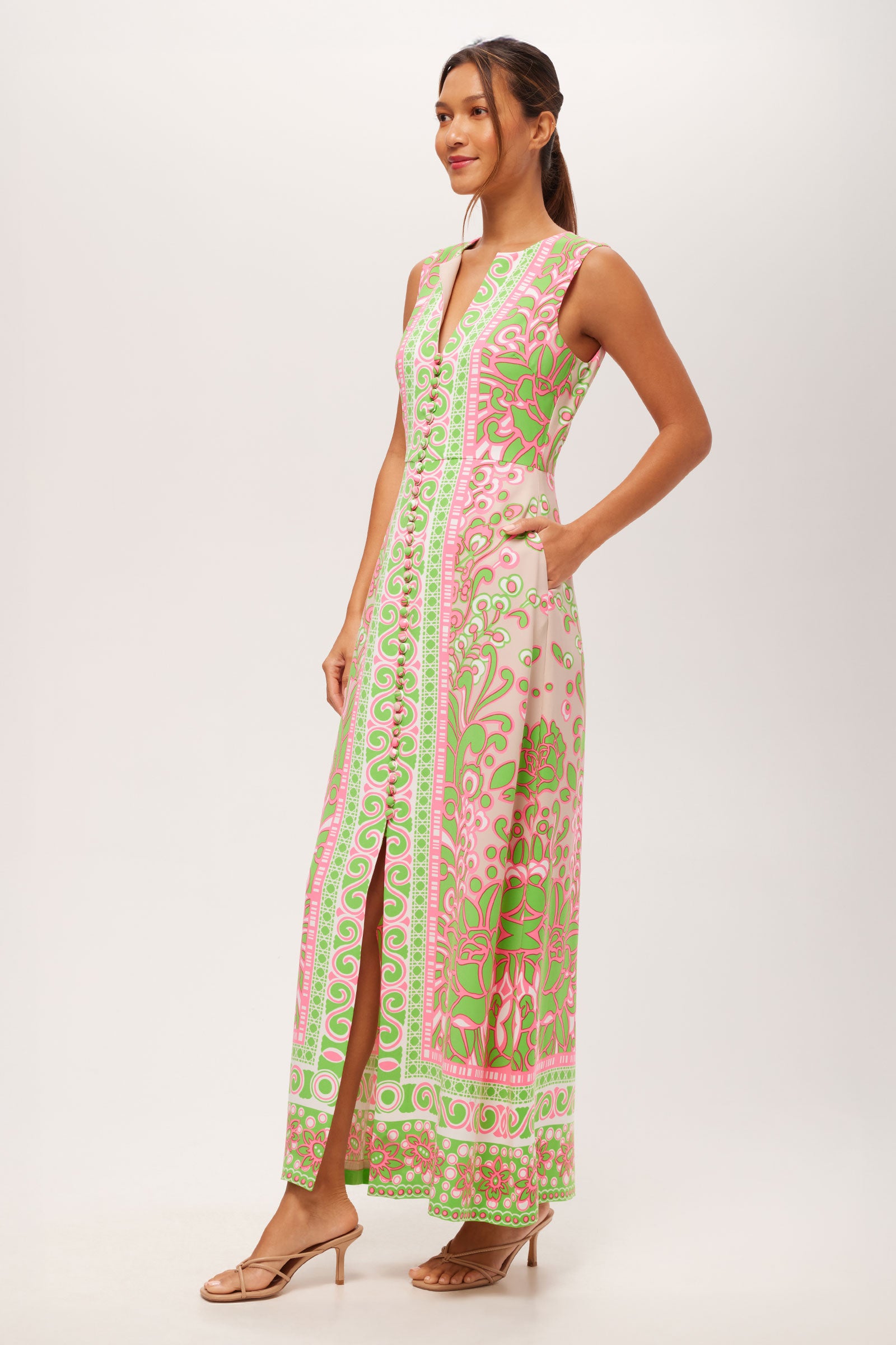 A woman smiles in the Novak Sleeveless Midi Dress - Petra Floral, a V-neck, sleeveless midi with pink, green, and white florals, front slit, and pockets. She pairs it with nude sandals and has her hair tied back.