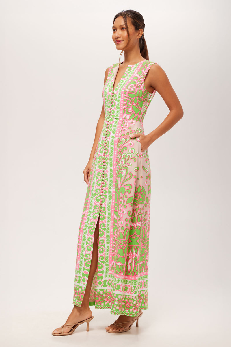 A woman smiles in the Novak Sleeveless Midi Dress - Petra Floral, a V-neck, sleeveless midi with pink, green, and white florals, front slit, and pockets. She pairs it with nude sandals and has her hair tied back.