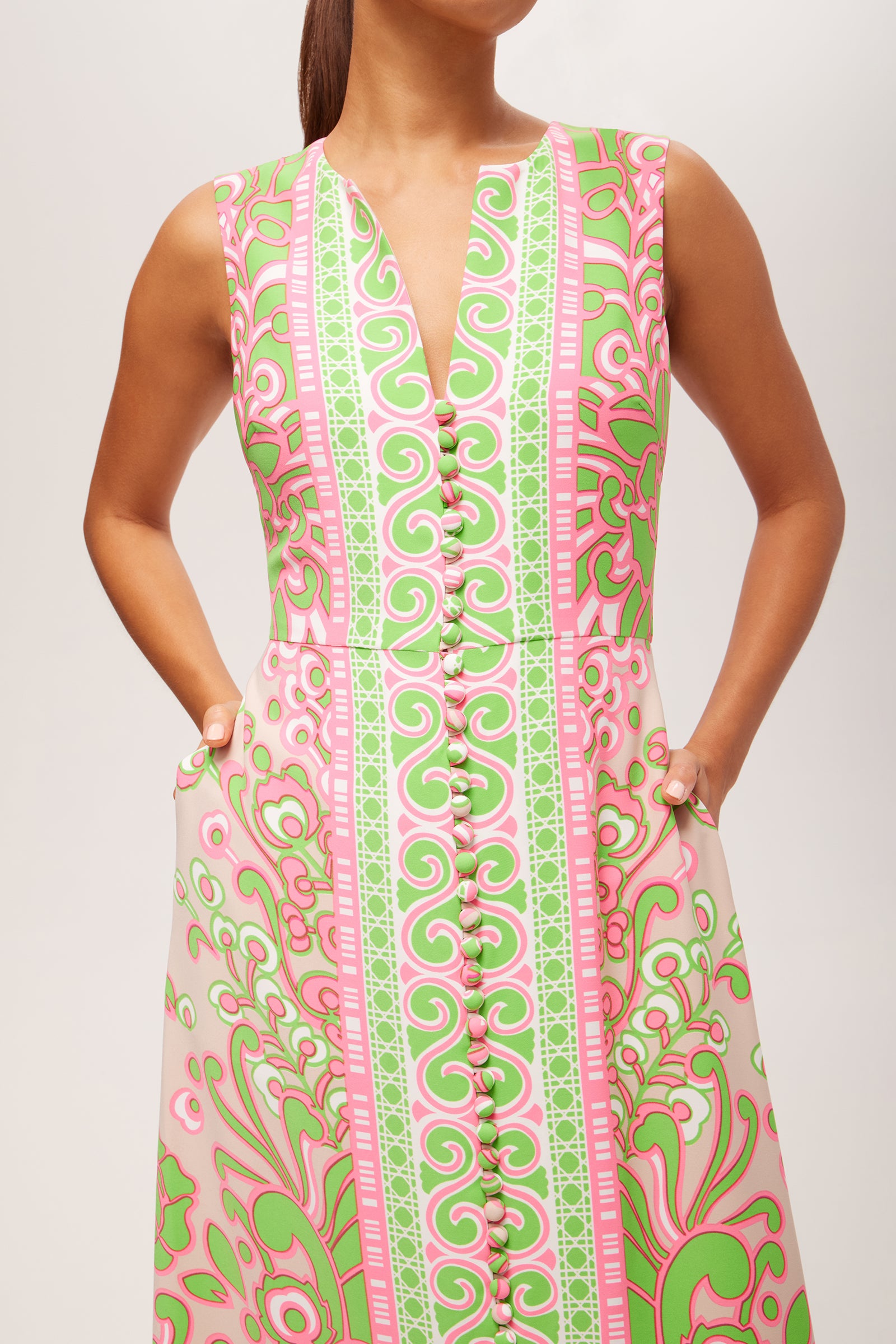 A woman wears the Novak Sleeveless Midi Dress - Petra Floral, featuring a deep V-neck, front buttons, pockets, and a pink, green, and white floral print. Her hands rest in her pockets as she turns her head slightly to the side.