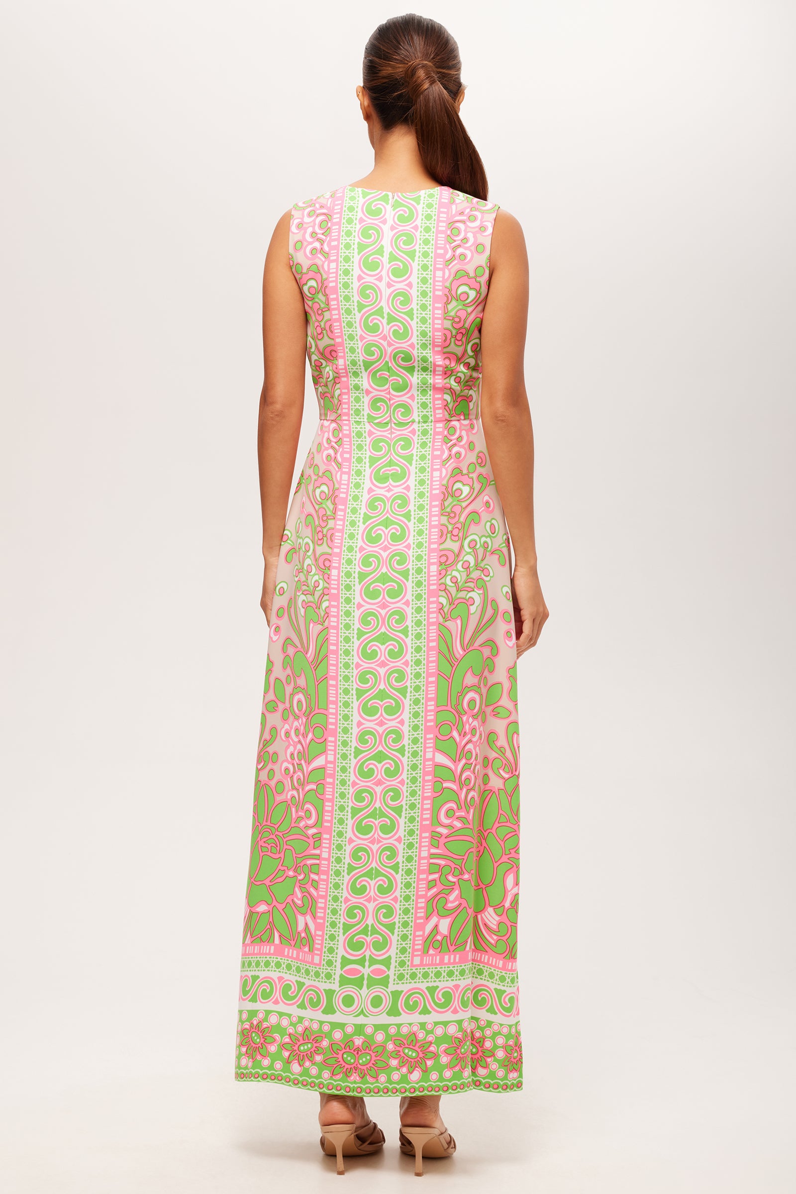 A woman with brown hair in a ponytail stands with her back to the camera against a plain light background, wearing the Novak Sleeveless Midi Dress - Petra Floral, featuring pink, green, and white ornate patterns.