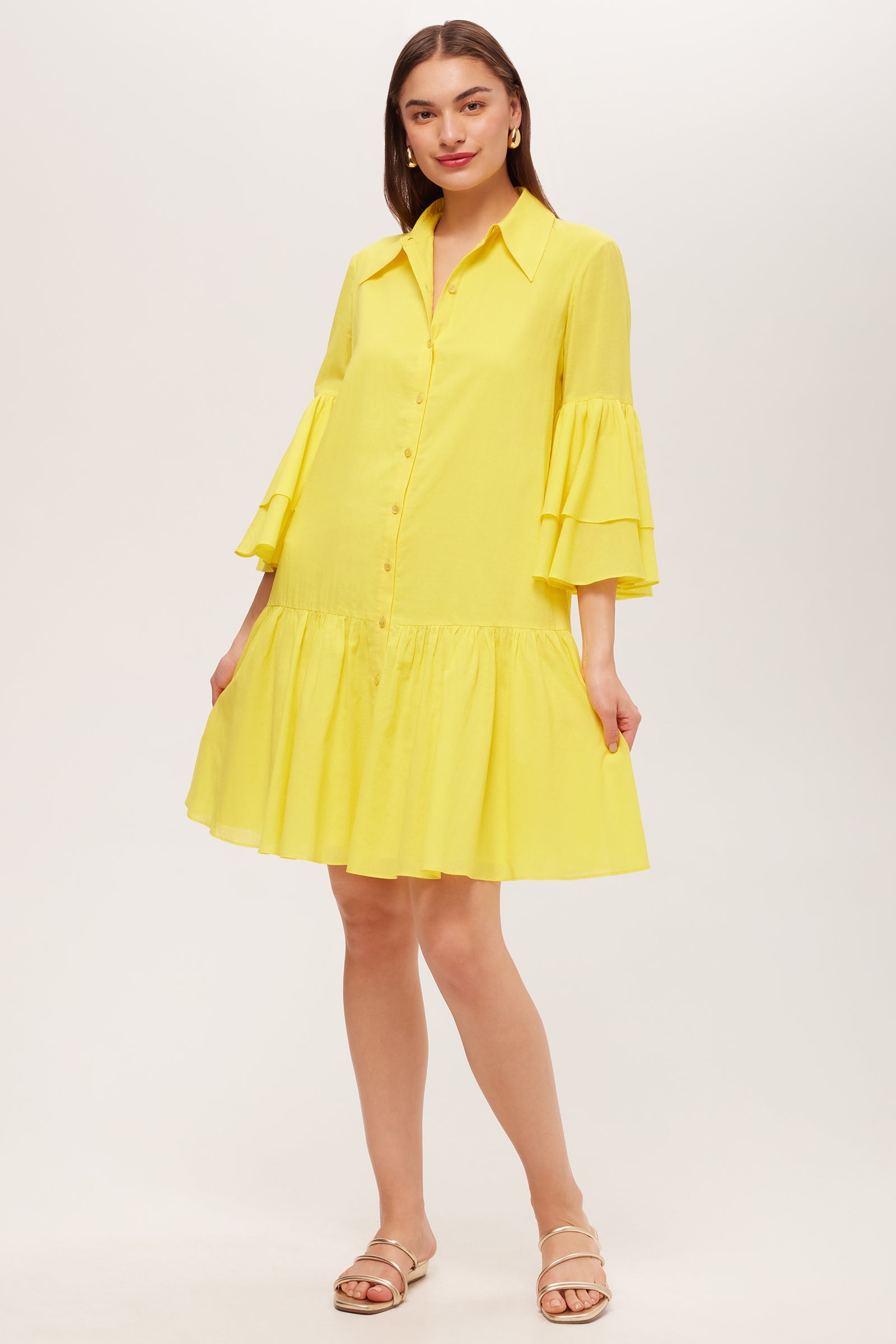 A woman wears the Bounce 3/4-Sleeve Ruffle Shirtdress, a bright yellow, loose-fit dress with tiered ruffle sleeves, a flared ruffled hem, front buttons, and a collar. She pairs it with flat sandals.
