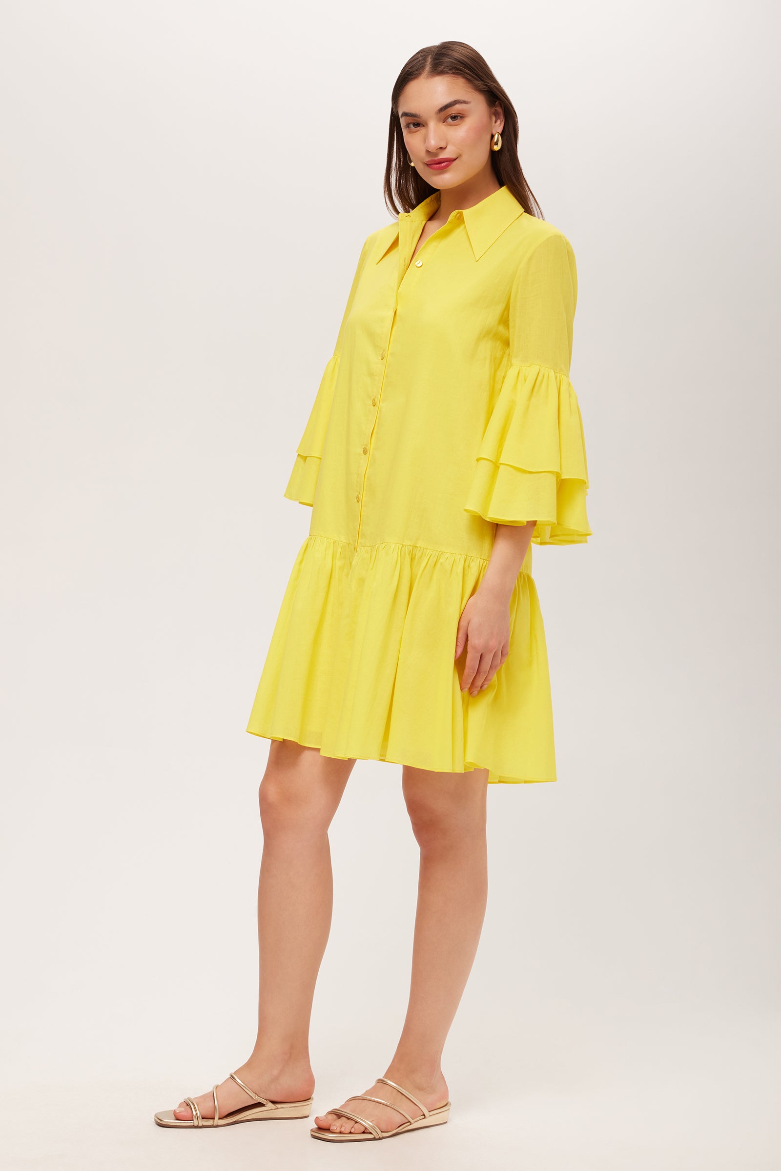 A woman with straight brown hair wears the Bounce 3/4-Sleeve Ruffle Shirtdress in Desert Sun, featuring tiered ruffled sleeves and a flared skirt. She pairs it with flat, strappy sandals against a plain white background.