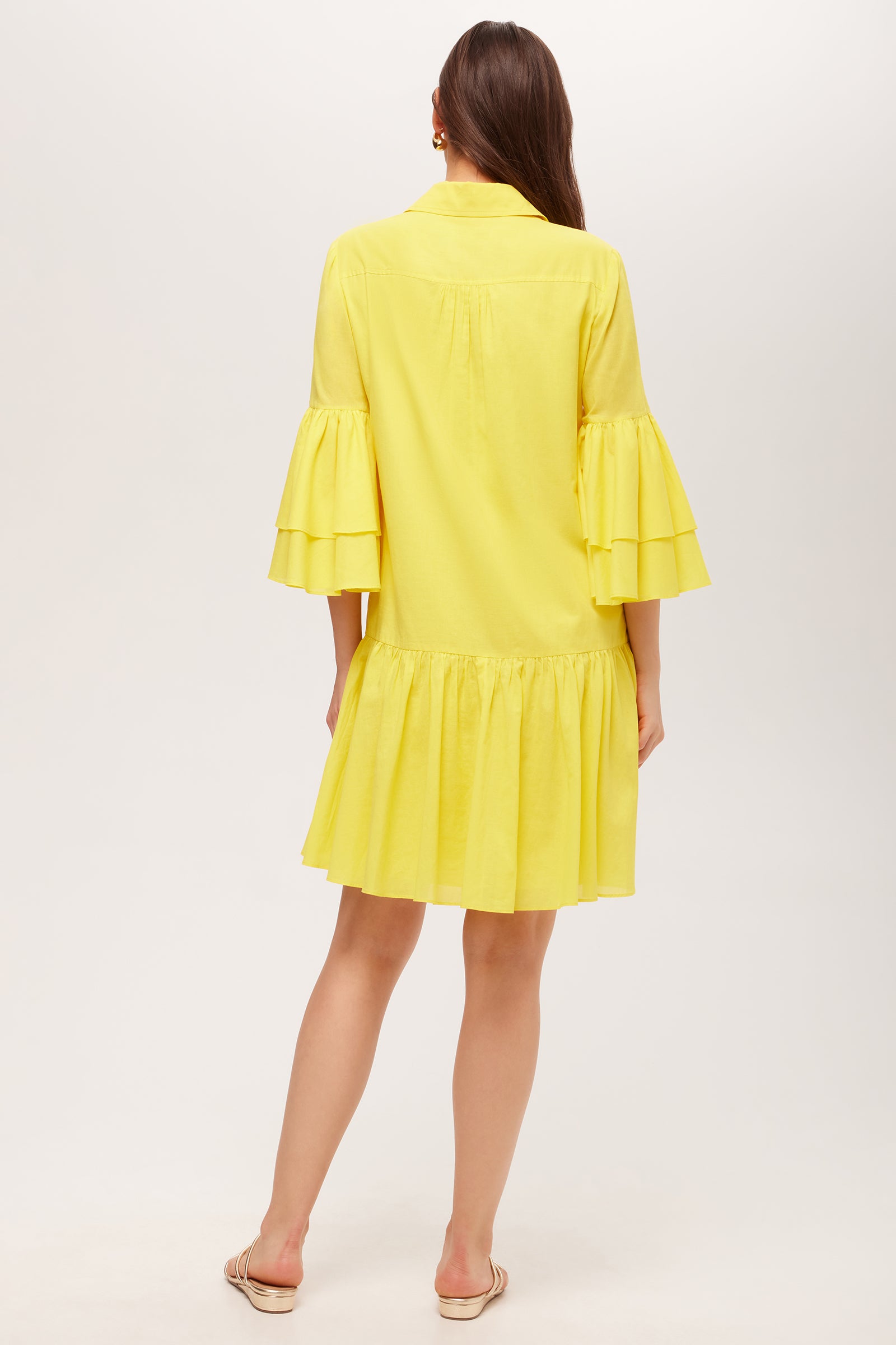 A woman with long brown hair faces away, wearing the Bounce 3/4-Sleeve Ruffle Shirtdress in yellow. The knee-length dress features loose fit, ruffled bell sleeves, and a ruffled hem. She wears flat shoes against a plain white background.