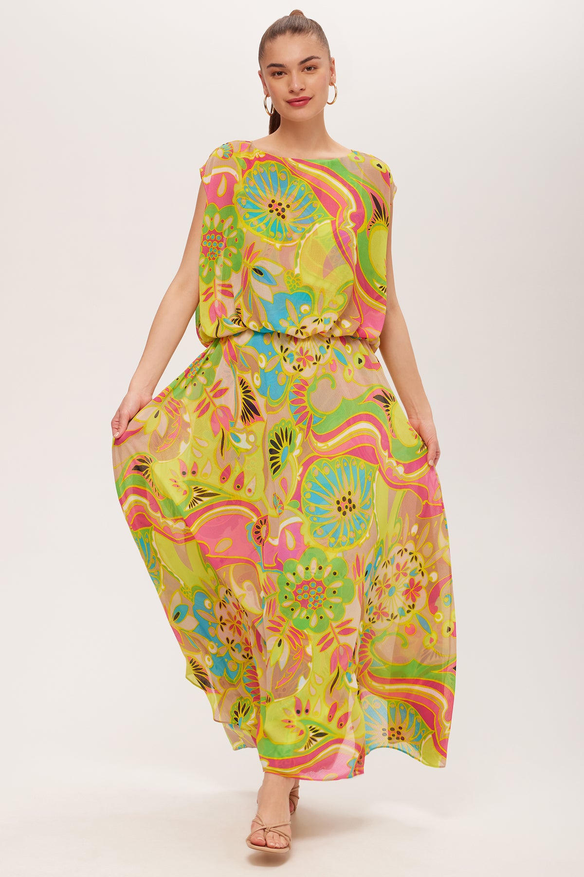 A woman stands against a plain light background, holding out the sides of her Vantage Short-Sleeve Maxi Dress - Venus, which features a flowing silhouette and a bright abstract floral pattern in yellow, green, pink, and blue.