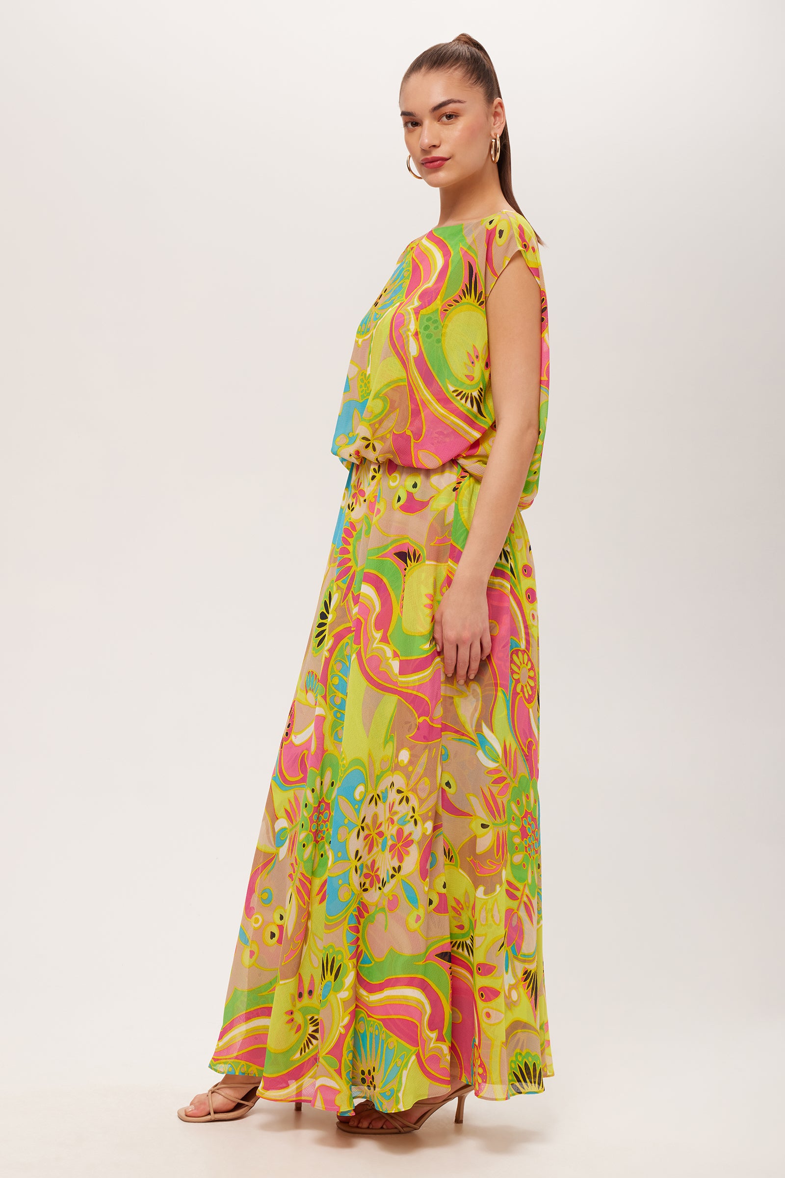 A woman stands sideways wearing the Vantage Short-Sleeve Maxi Dress - Venus, a colorful, floor-length dress with vibrant green, pink, yellow, and blue floral and abstract prints, paired with strappy heels against a plain white background.