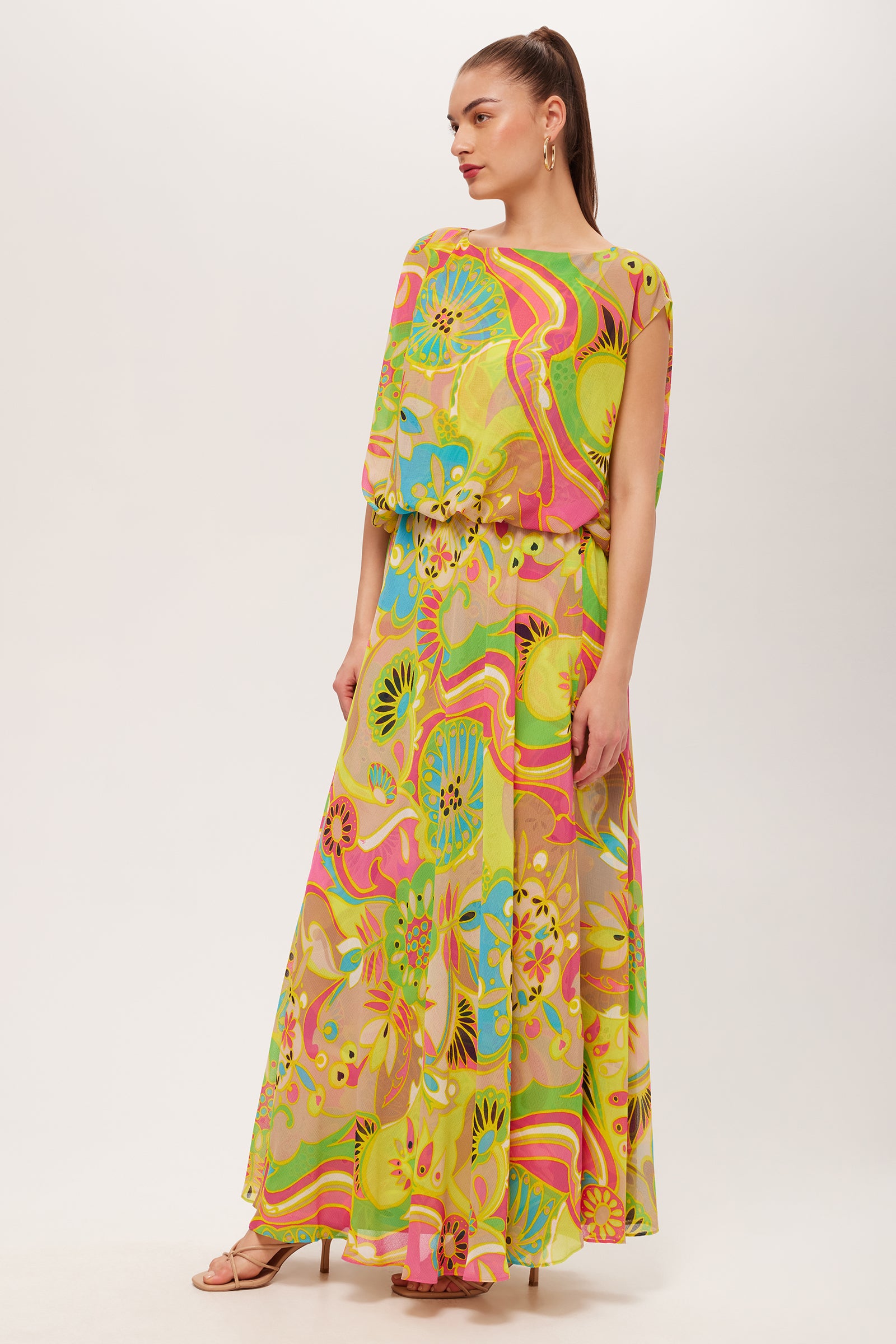 A woman wears the Vantage Short-Sleeve Maxi Dress in Venus, featuring a vibrant abstract floral pattern. Her hair is tied back, she accessorizes with hoop earrings and open-toe heels, standing against a plain white background.