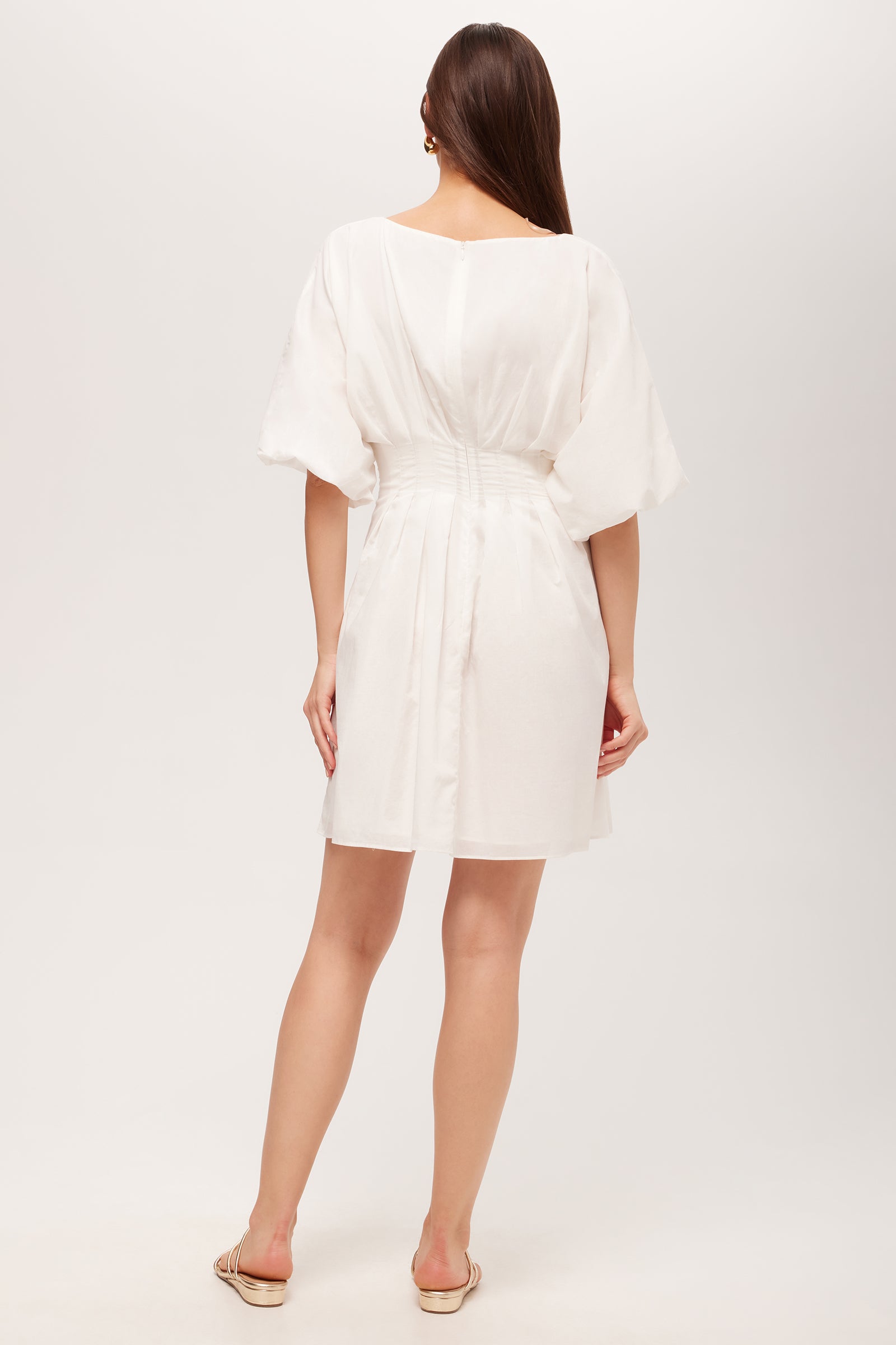 A woman stands with her back to the camera, wearing the Gian 2 Short-Sleeve Pleated Dress—short, white, featuring loose sleeves and a cinched waist—paired with flat beige sandals against a plain light background.