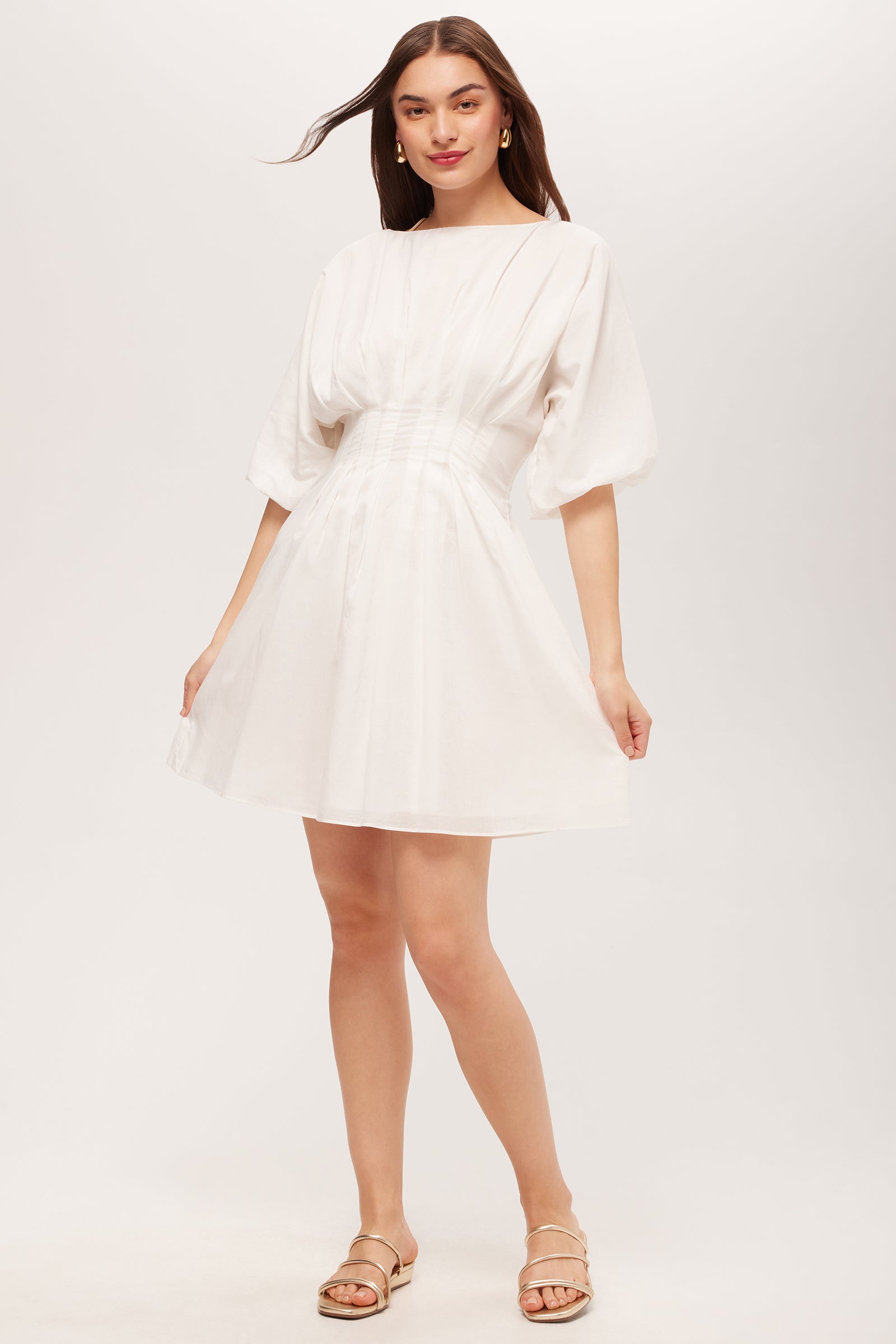 A woman wearing the Gian 2 Short-Sleeve Pleated Dress in white stands against a plain background. She has long hair, is smiling softly, and pairs the dress with flat sandals.