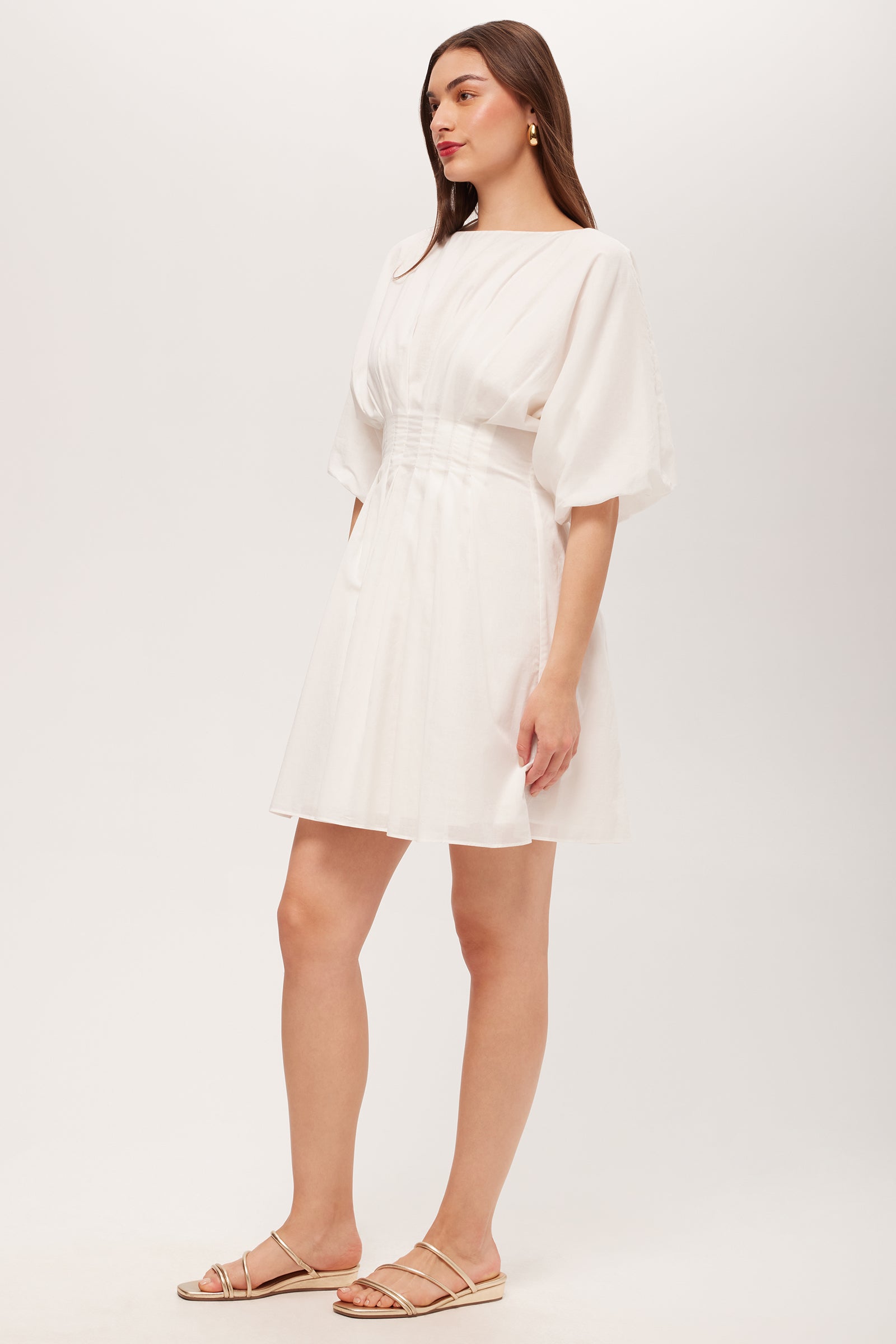 A woman with long brown hair poses sideways against a white background, wearing the Gian 2 Short-Sleeve Pleated Dress—short, white, with elbow-length puff sleeves and a belted waist—paired with beige flat sandals.