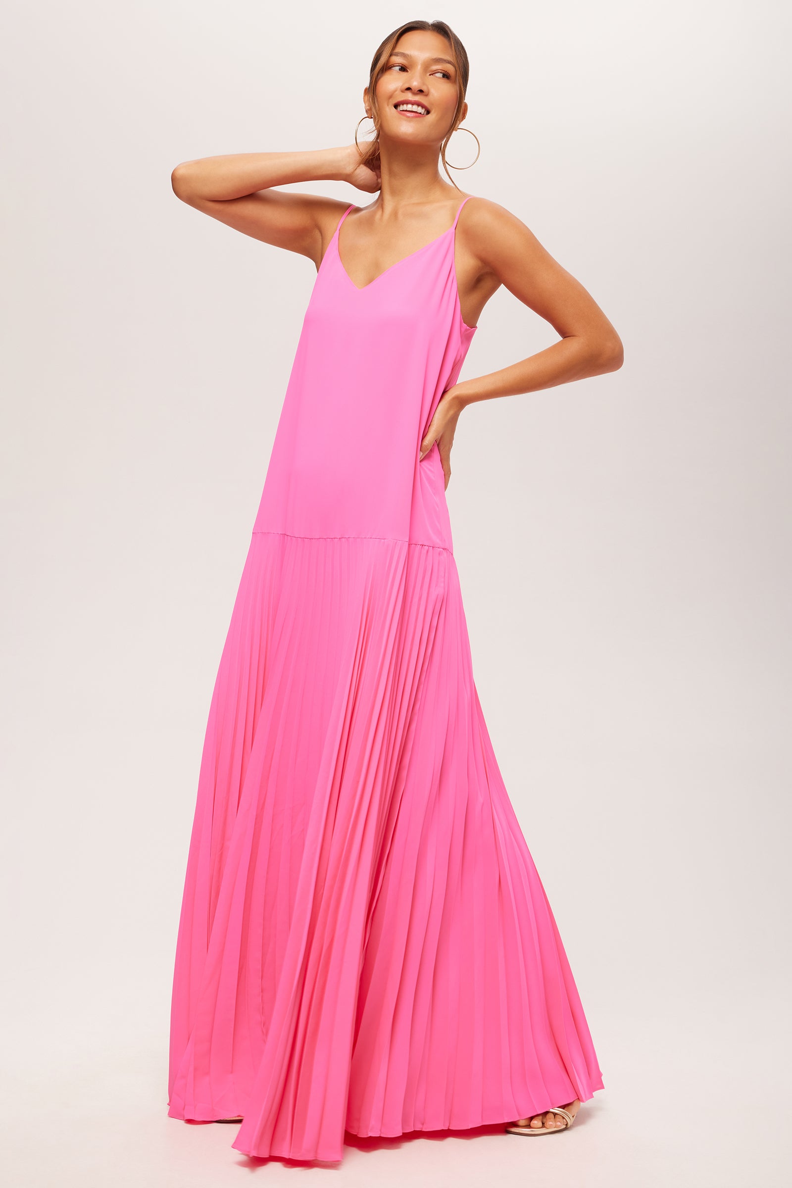 A woman stands smiling with one hand on her hip and the other on her head, wearing the Carve Sleeveless Drop-Waist Pleated Maxi Dress in bright pink, featuring a V-neckline and flowy maxi-length silhouette against a light background.
