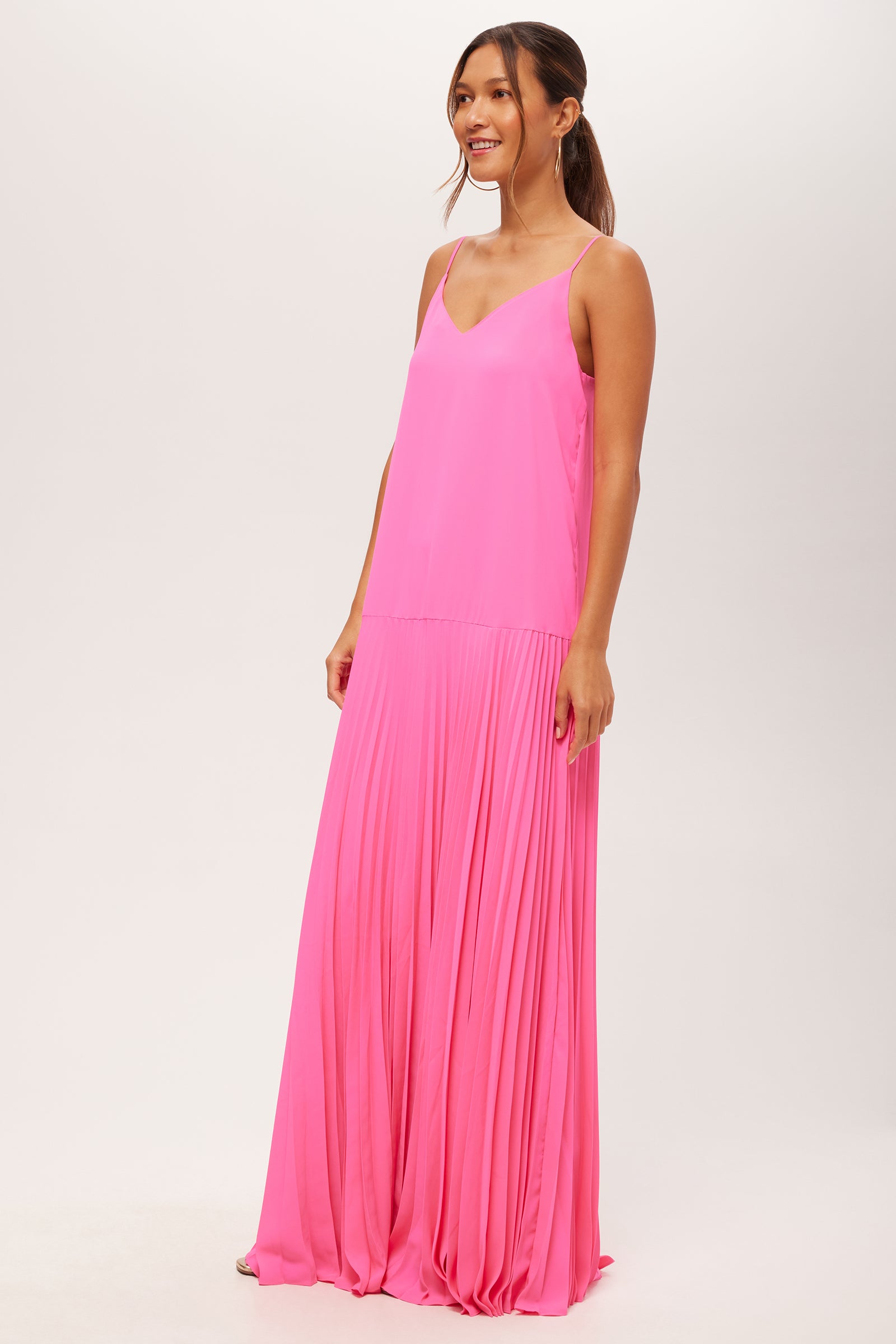 A woman smiles with her hair in a low ponytail, wearing the Carve Sleeveless Drop-Waist Pleated Maxi Dress—a bright pink, floor-length dress with a V-neckline—against a plain light background.