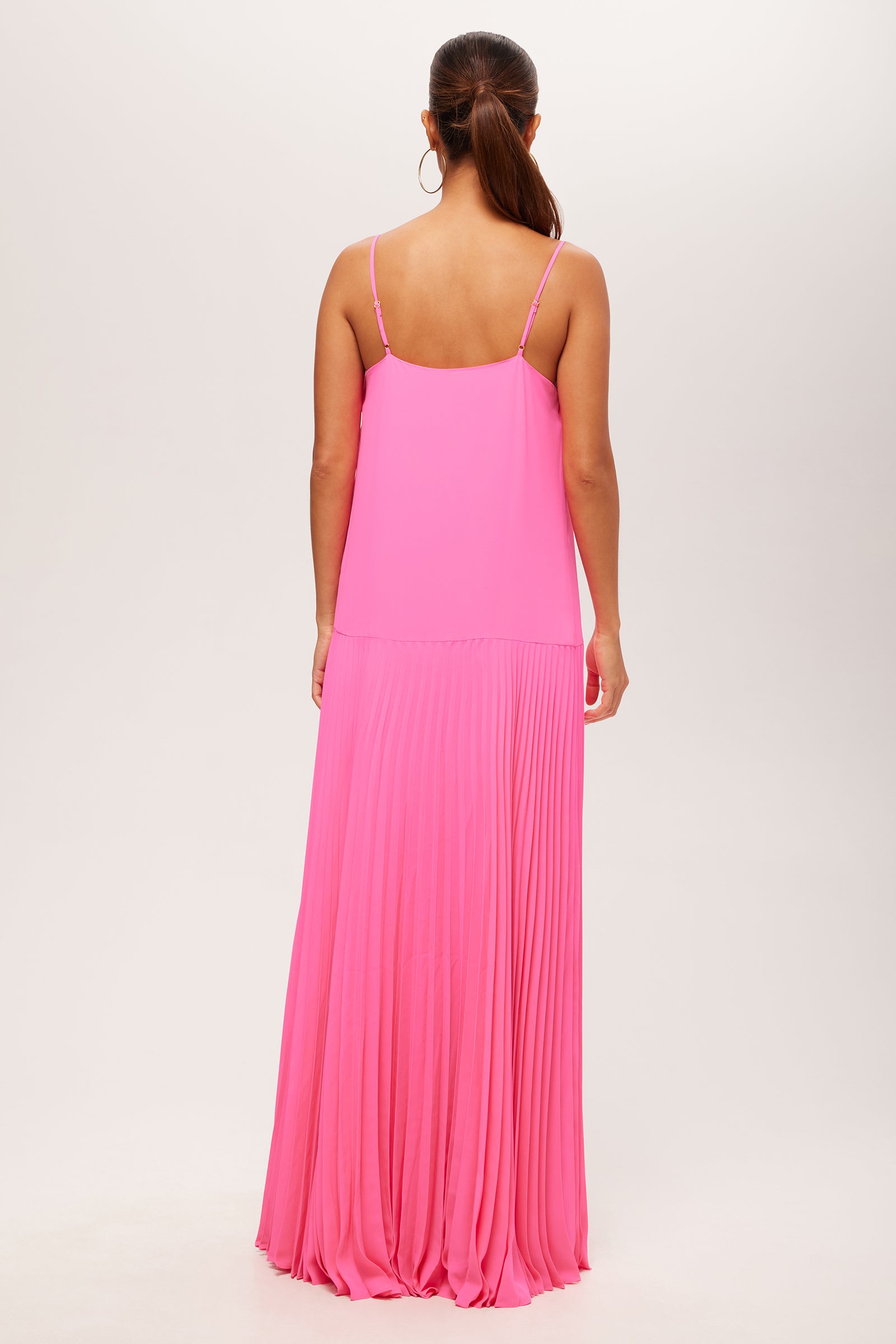 A woman with her hair in a low ponytail faces away, wearing the Carve Sleeveless Drop-Waist Pleated Maxi Dress in bright pink, featuring thin straps and a pleated skirt, set against a plain light background.