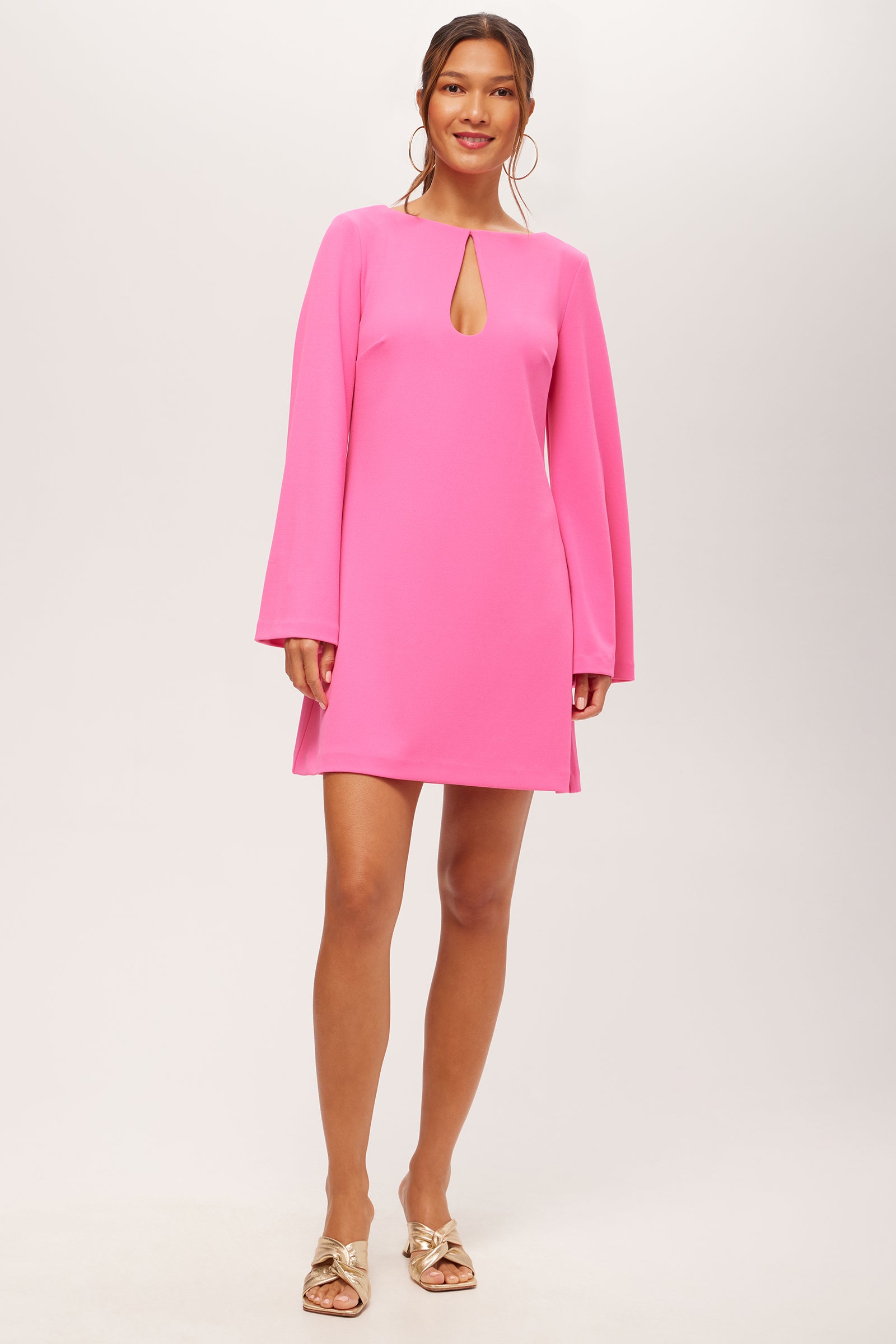 A woman stands against a light background wearing the Singles Long-Sleeve Keyhole Dress in Love All Pink, featuring a front keyhole detail. Her hair is tied back, and she accessorizes with large hoop earrings and gold open-toed sandals.