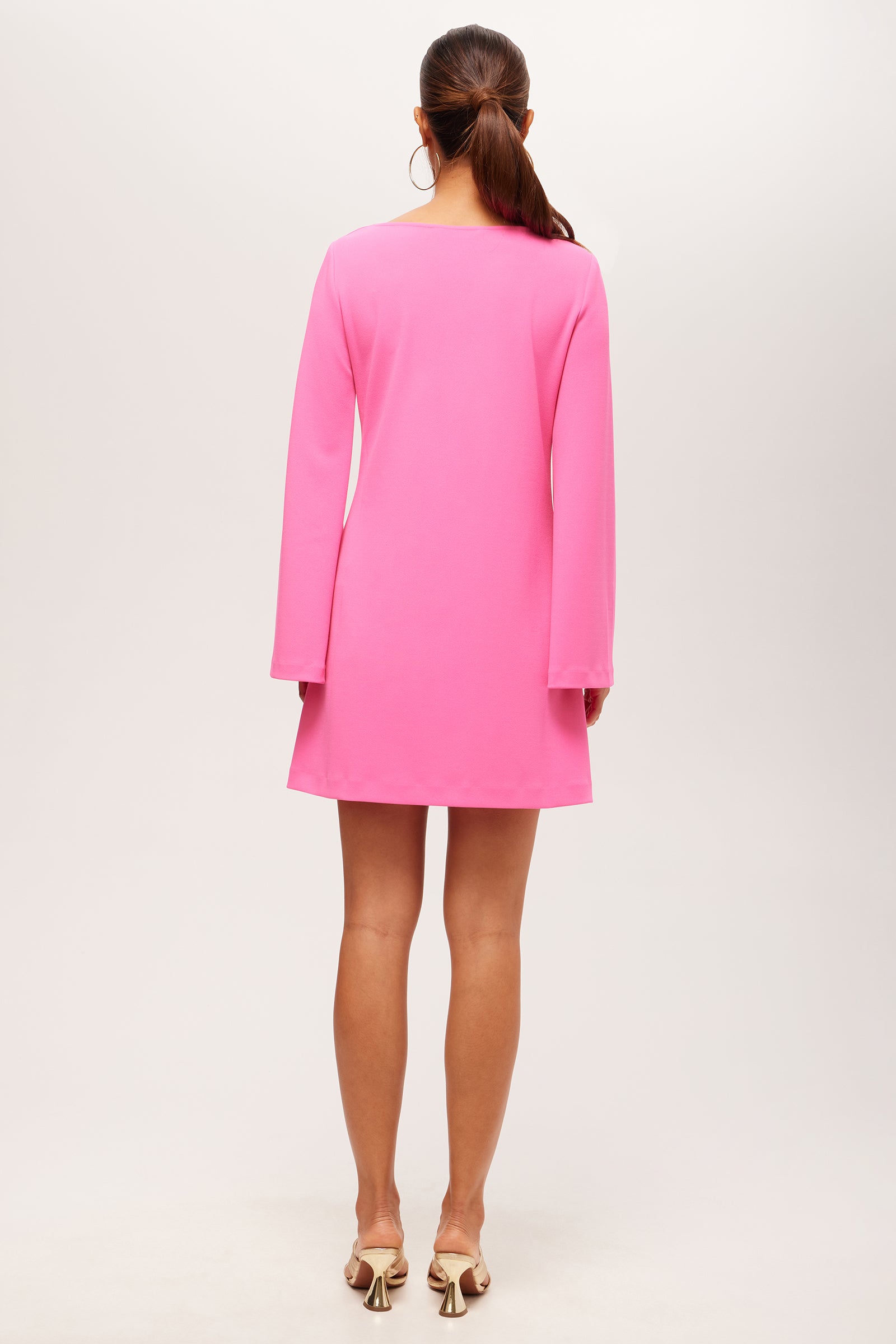 A woman stands facing away in the Singles Long-Sleeve Keyhole Dress - Crepe Knit, a bright pink mini dress with long sleeves. She pairs it with white heeled sandals, her brown hair tied back against a plain light background.