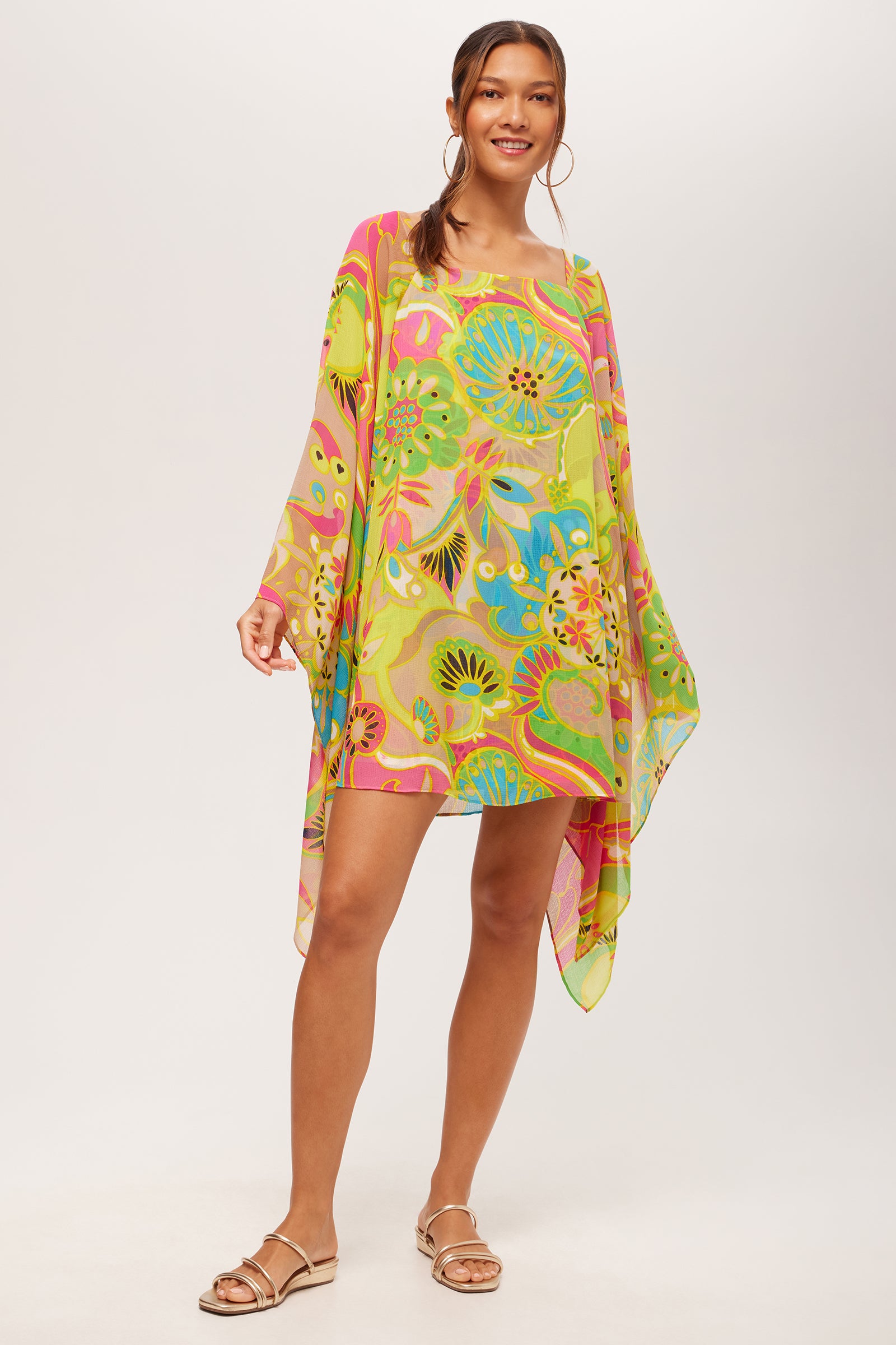 A woman smiles in the Aqualane Side Flounce Dress - Venus, a loose, colorful floral piece with long, wide sleeves. She completes the look with tan flat sandals and gold hoop earrings against a plain light background.