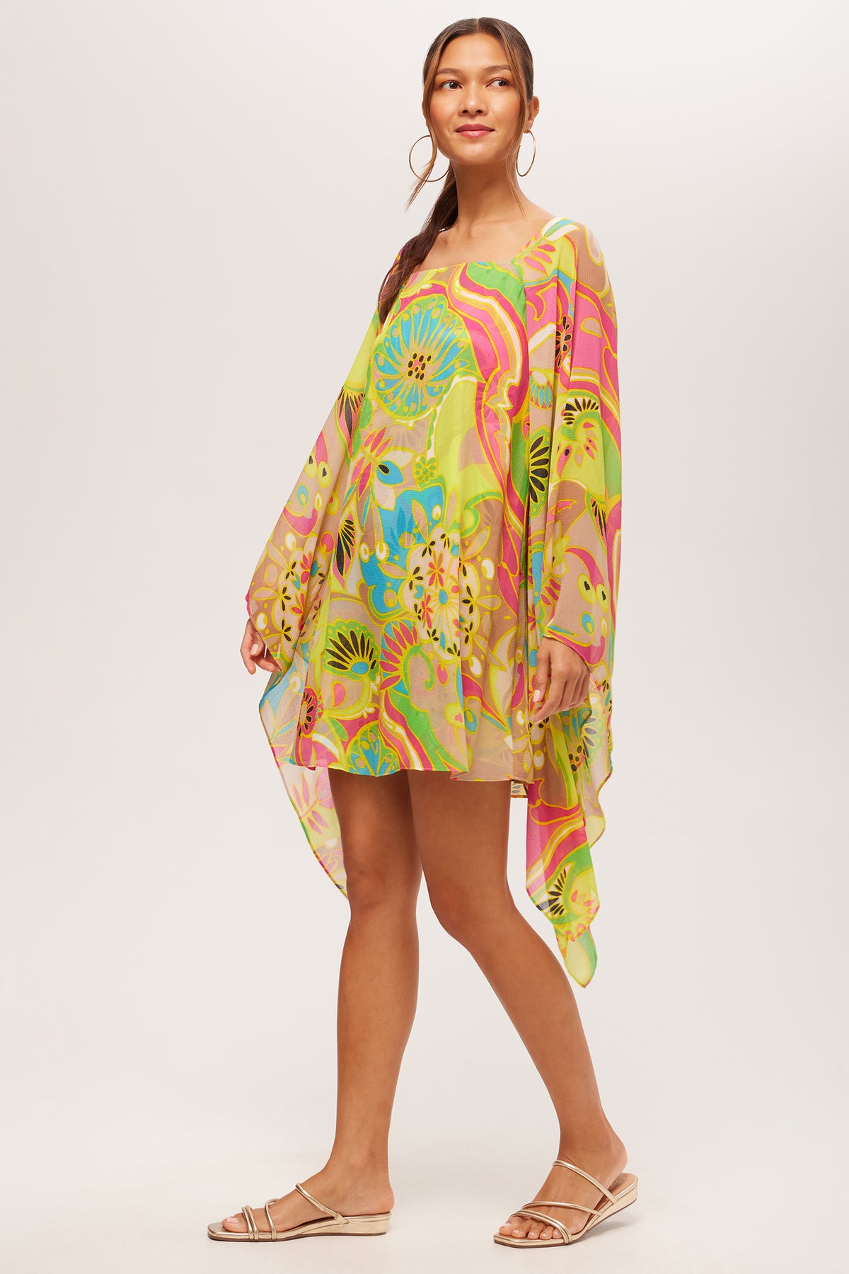 A woman smiles slightly in the Aqualane Side Flounce Dress by Venus—a lightweight, long-sleeve dress featuring an asymmetrical cut and a colorful floral and abstract pattern in green, yellow, blue, and pink. She pairs it with flat sandals and hoop earrings.