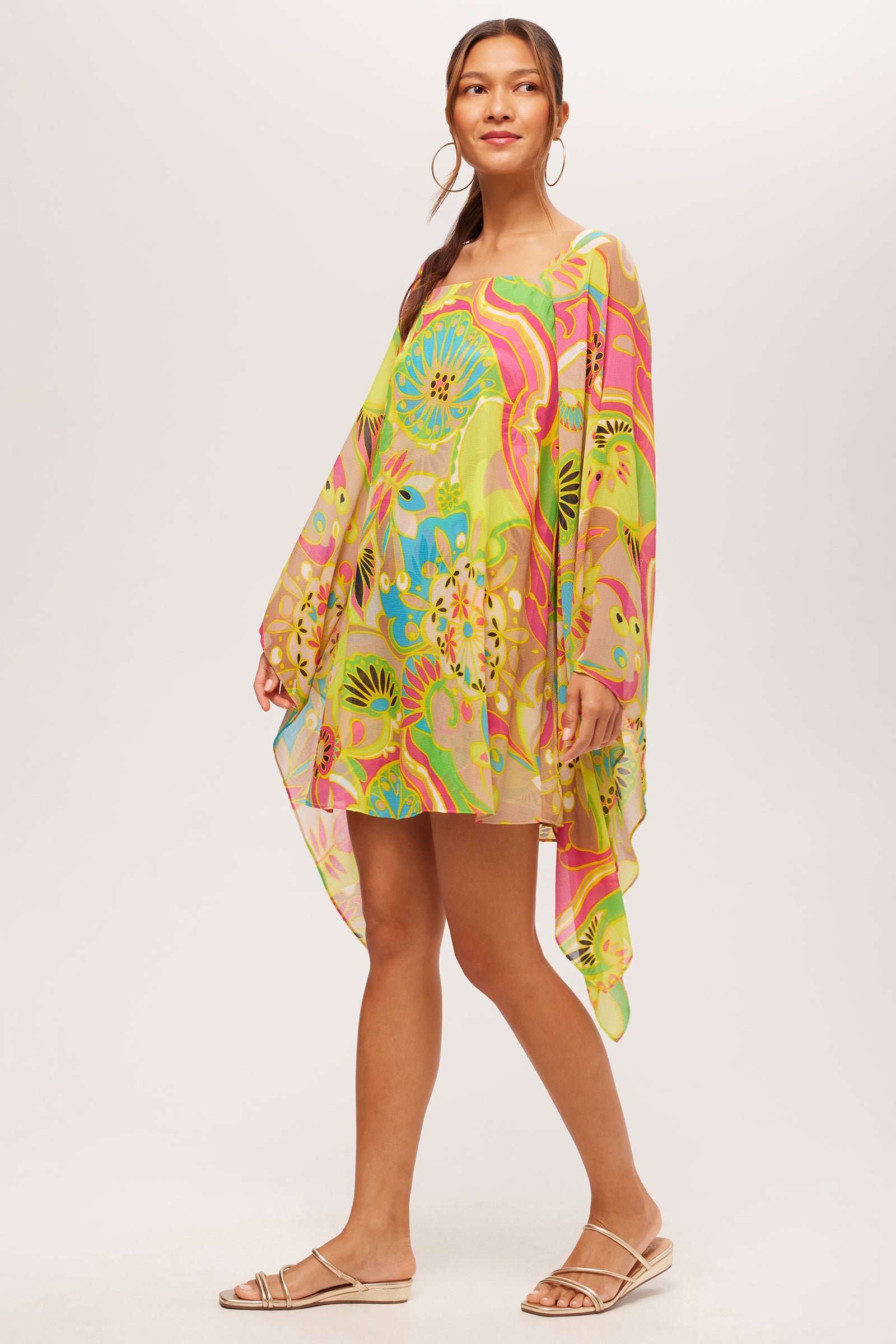 A woman smiles slightly in the Aqualane Side Flounce Dress by Venus—a lightweight, long-sleeve dress featuring an asymmetrical cut and a colorful floral and abstract pattern in green, yellow, blue, and pink. She pairs it with flat sandals and hoop earrings.