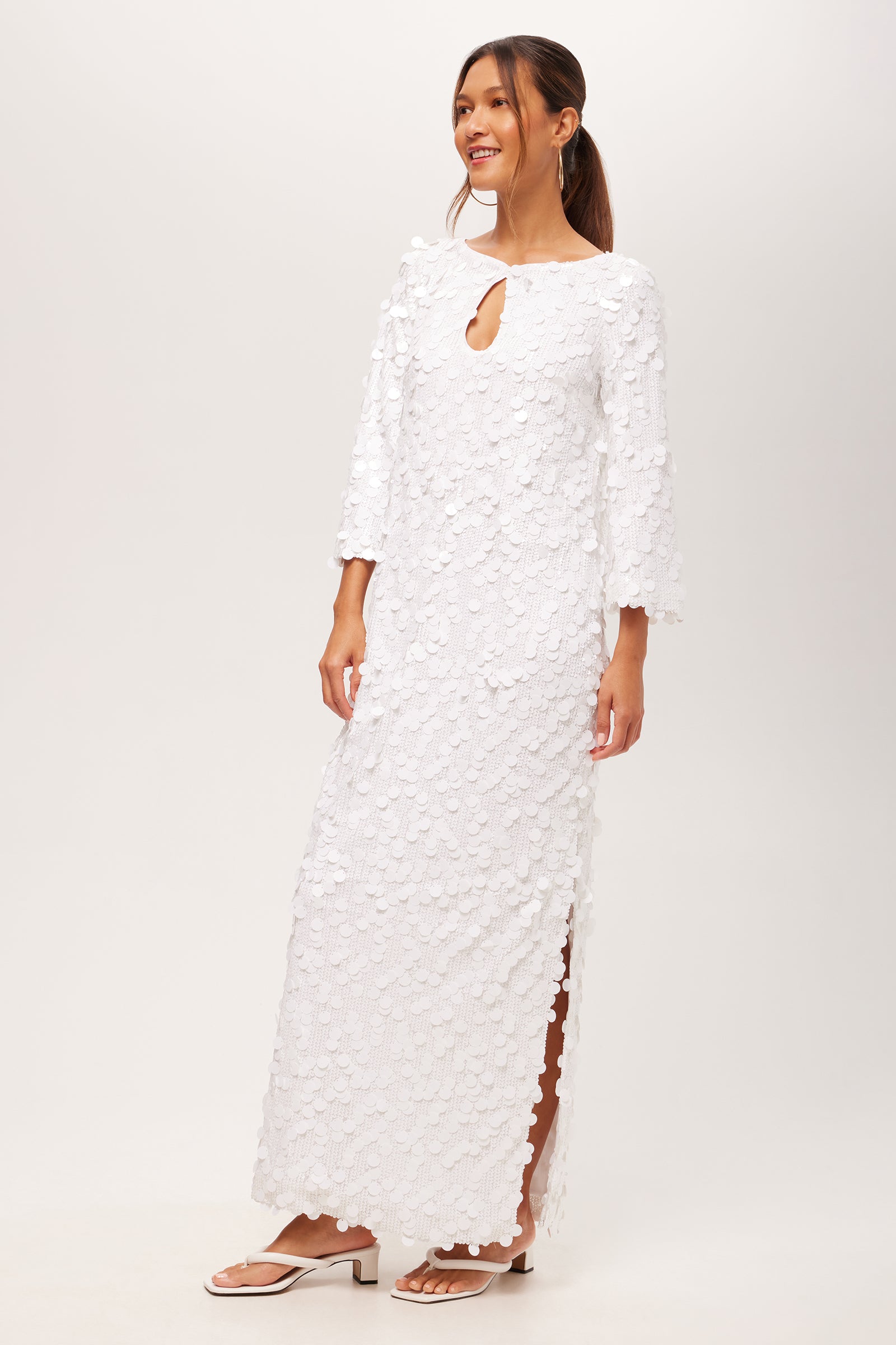 A woman stands smiling against a plain background, wearing the Singles 3/4-Sleeve Keyhole Maxi Dress—a long white textured dress with three-quarter sleeves, keyhole neckline, side slit—and white sandals. Her hair is styled in a low ponytail.