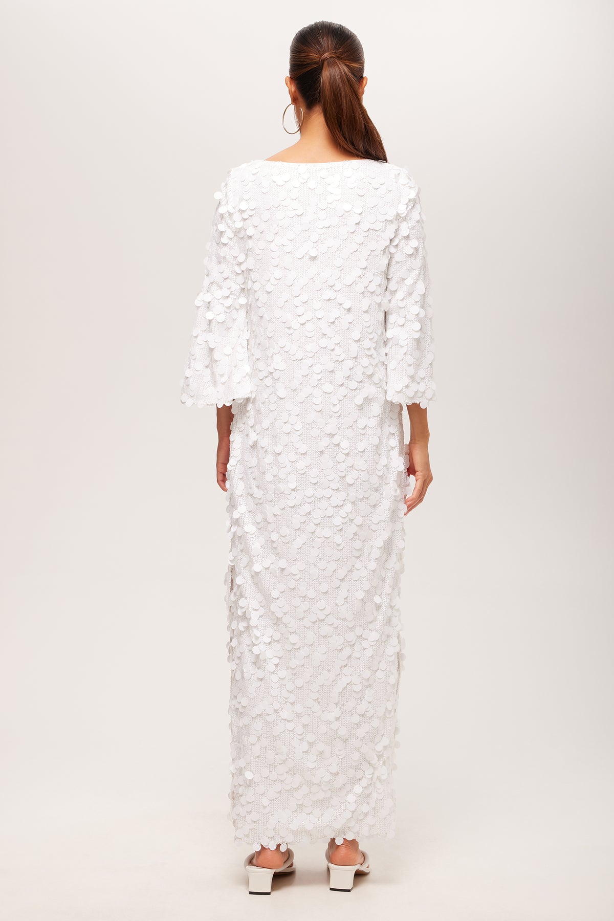 A woman with long brown hair in a ponytail faces away, wearing the Singles 3/4-Sleeve Keyhole Maxi Dress—an ankle-length white dress with textured petal details and wide sleeves—paired with white block-heeled shoes.