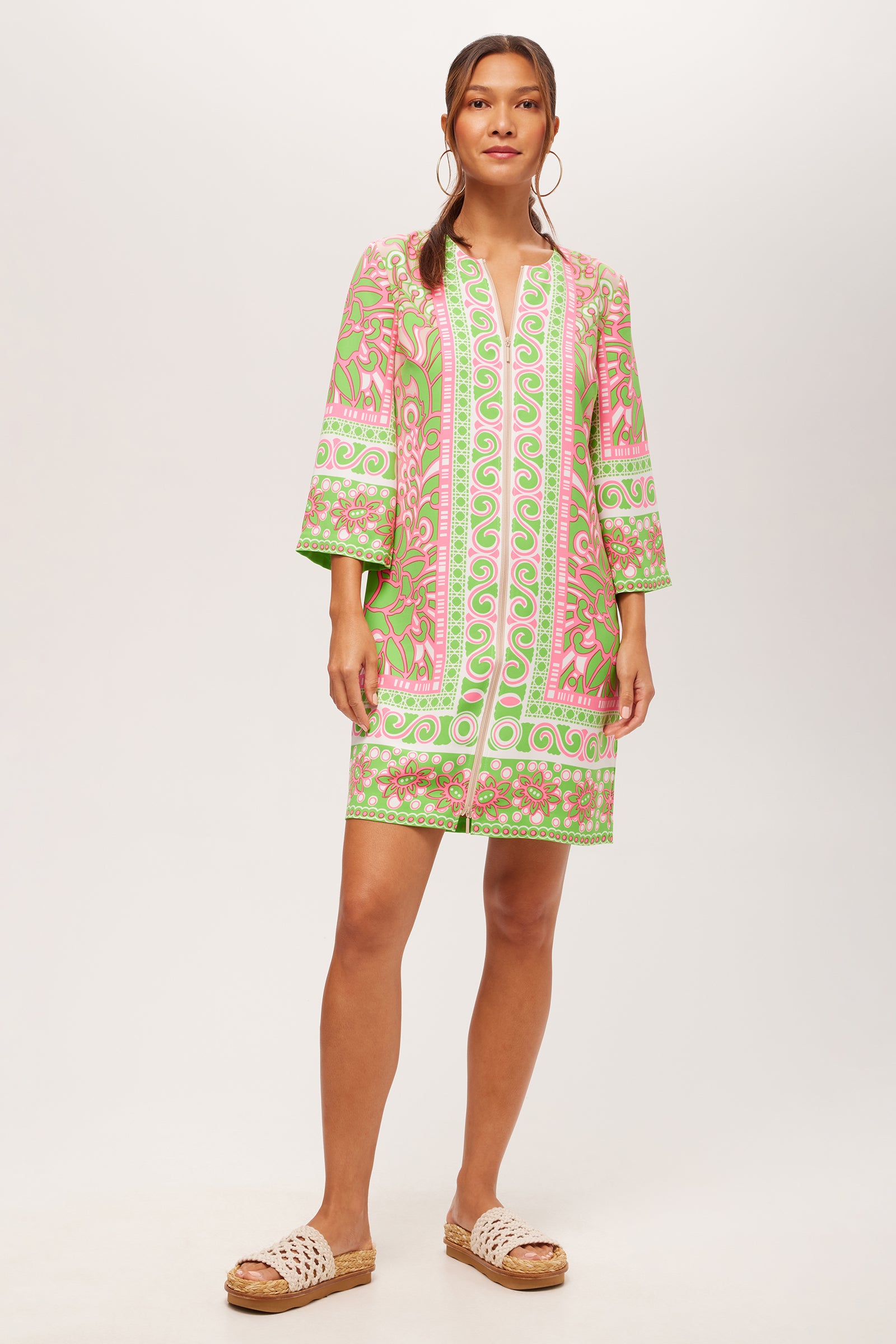 A woman in the Botanical 3/4-Sleeve Zip Dress in Petra Floral stands against a plain white background, wearing hoop earrings, slide sandals, and her hair pulled back.