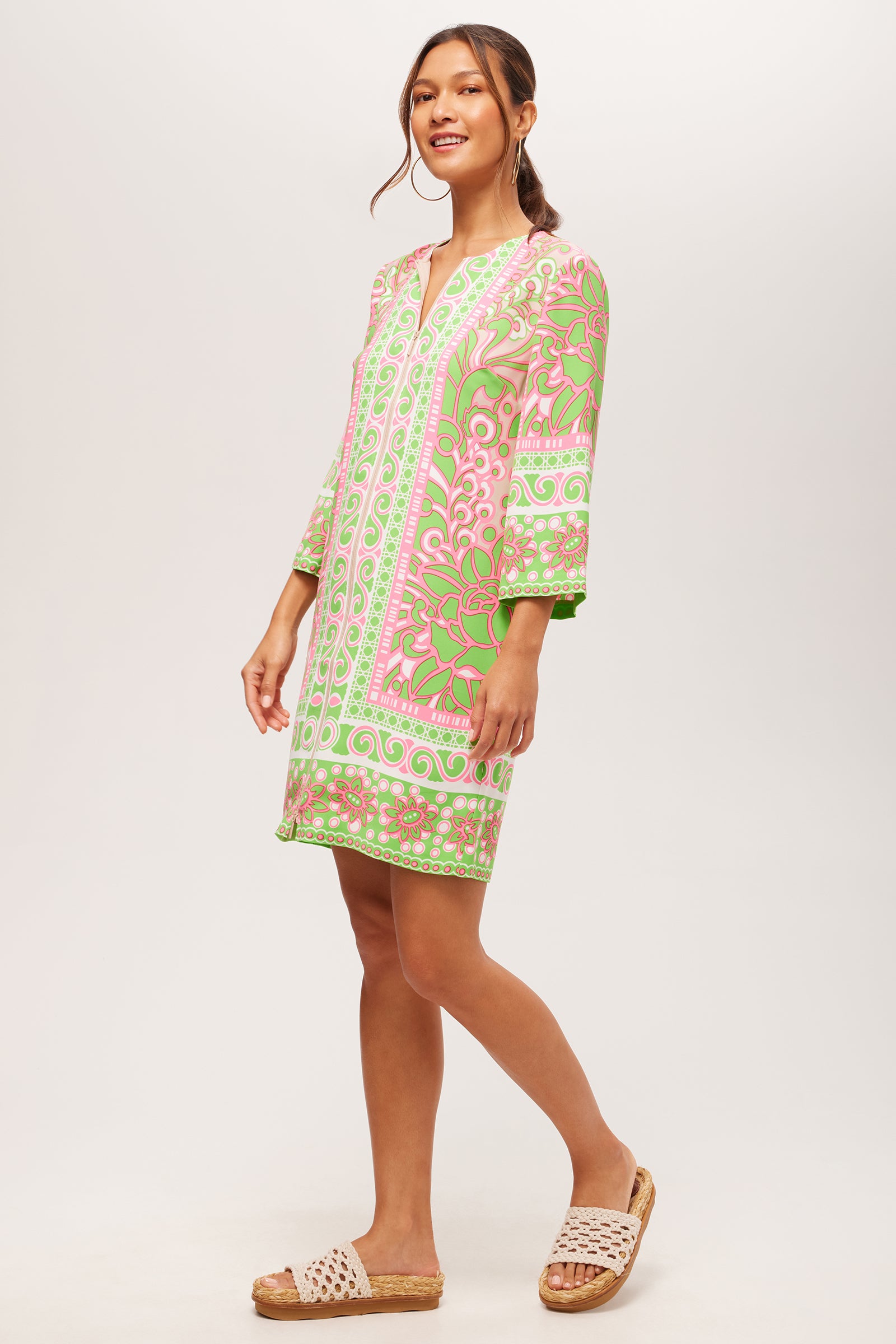 A woman stands smiling on a white background wearing the Botanical 3/4-Sleeve Zip Dress - Petra Floral, a pink and green patterned knee-length dress, paired with light-colored woven slide sandals.